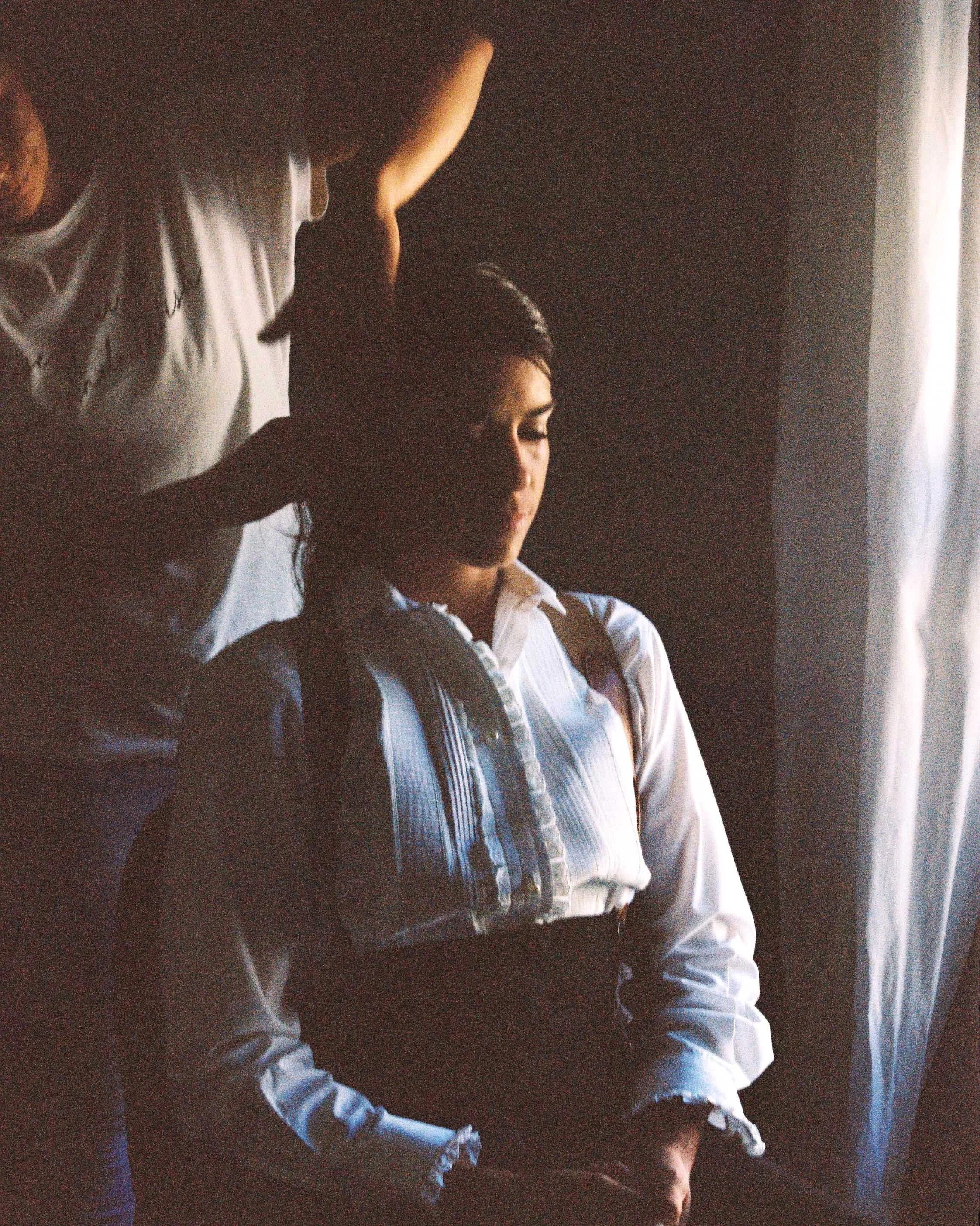 A young woman with closed eyes, wearing a white blouse, sits with a backpack on her shoulders next to a window with light coming through. A person standing behind her appears to be adjusting her hair.
