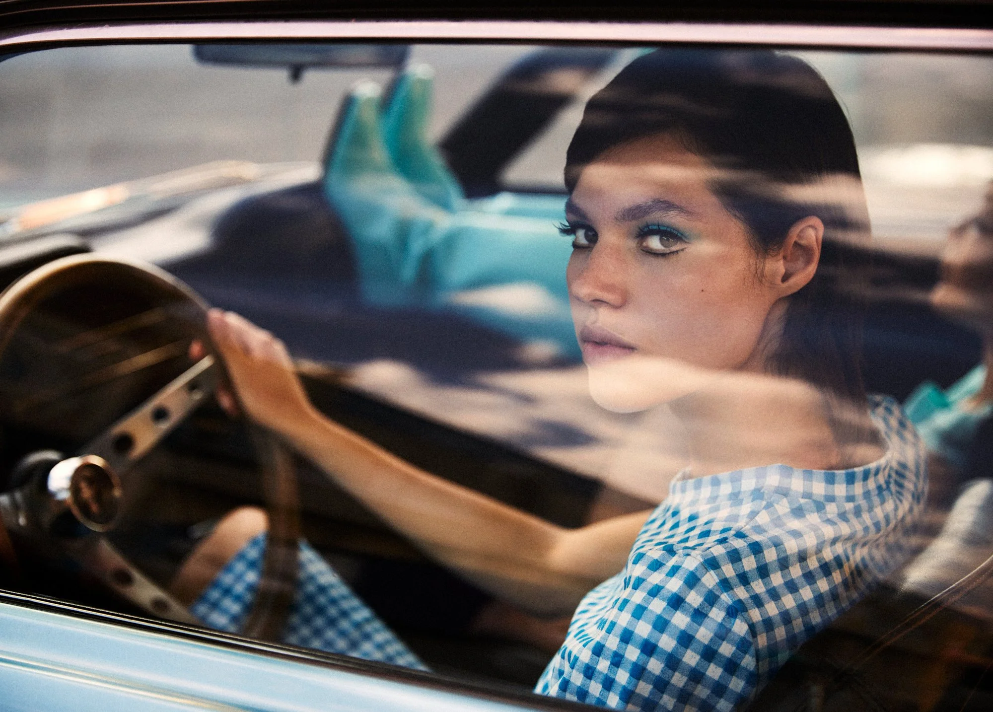 Woman with short dark hair and blue makeup sitting in car, looking through the windshield.