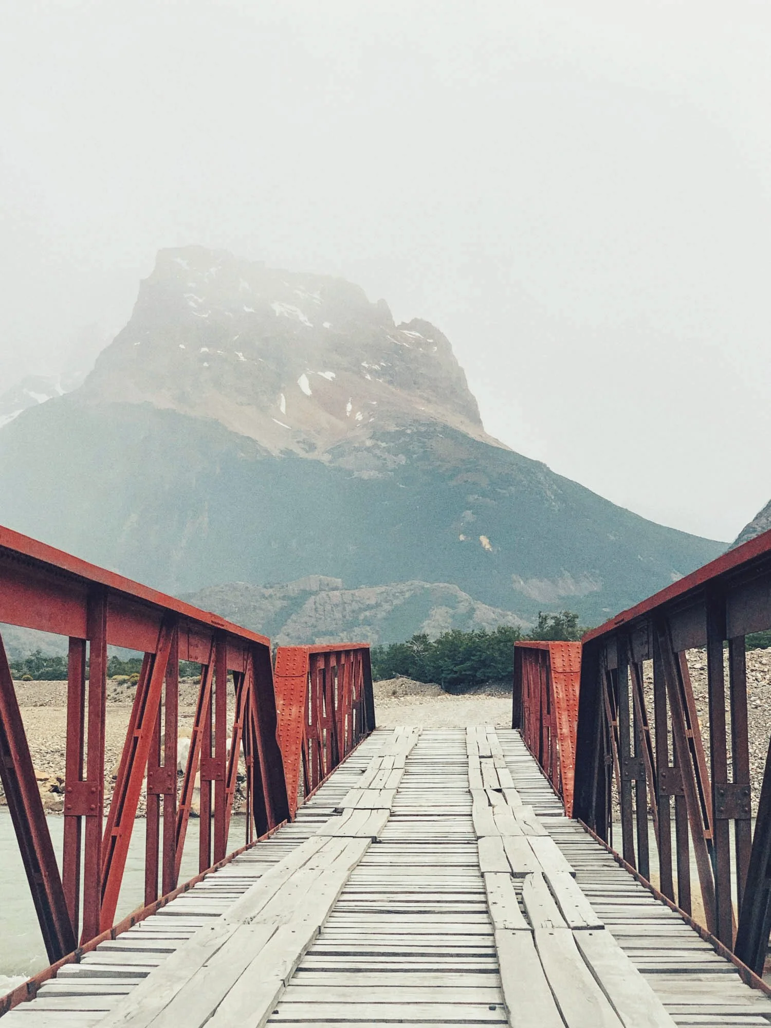 A wooden bridge with red railings leading towards a mountain in the distance, partly obscured by mist or fog.