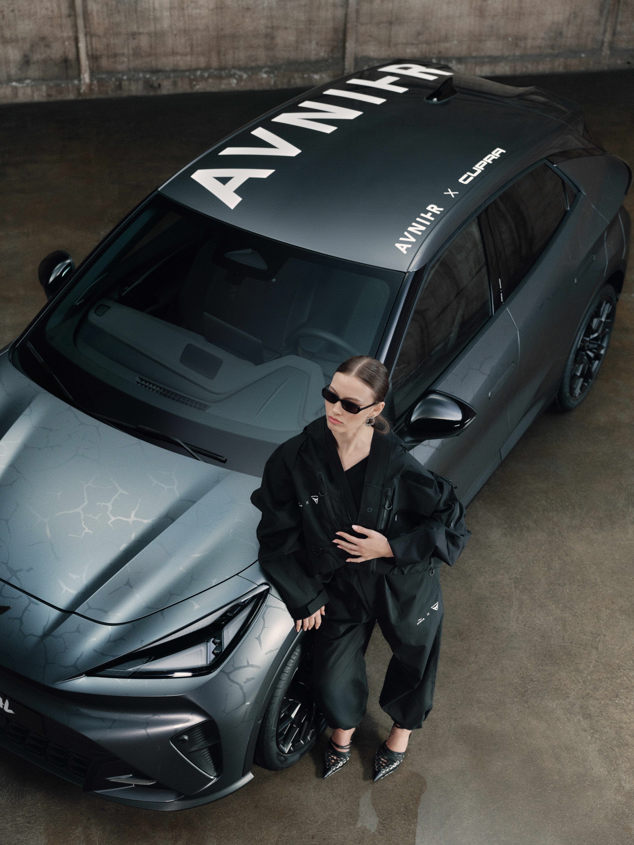 A woman with dark hair, sunglasses, and fashionable heels standing next to a sleek, modern gray car with a custom honeycomb pattern on the hood. The car has branding on the windshield and roof, including 'AVNIAR' and 'CUPRA'.