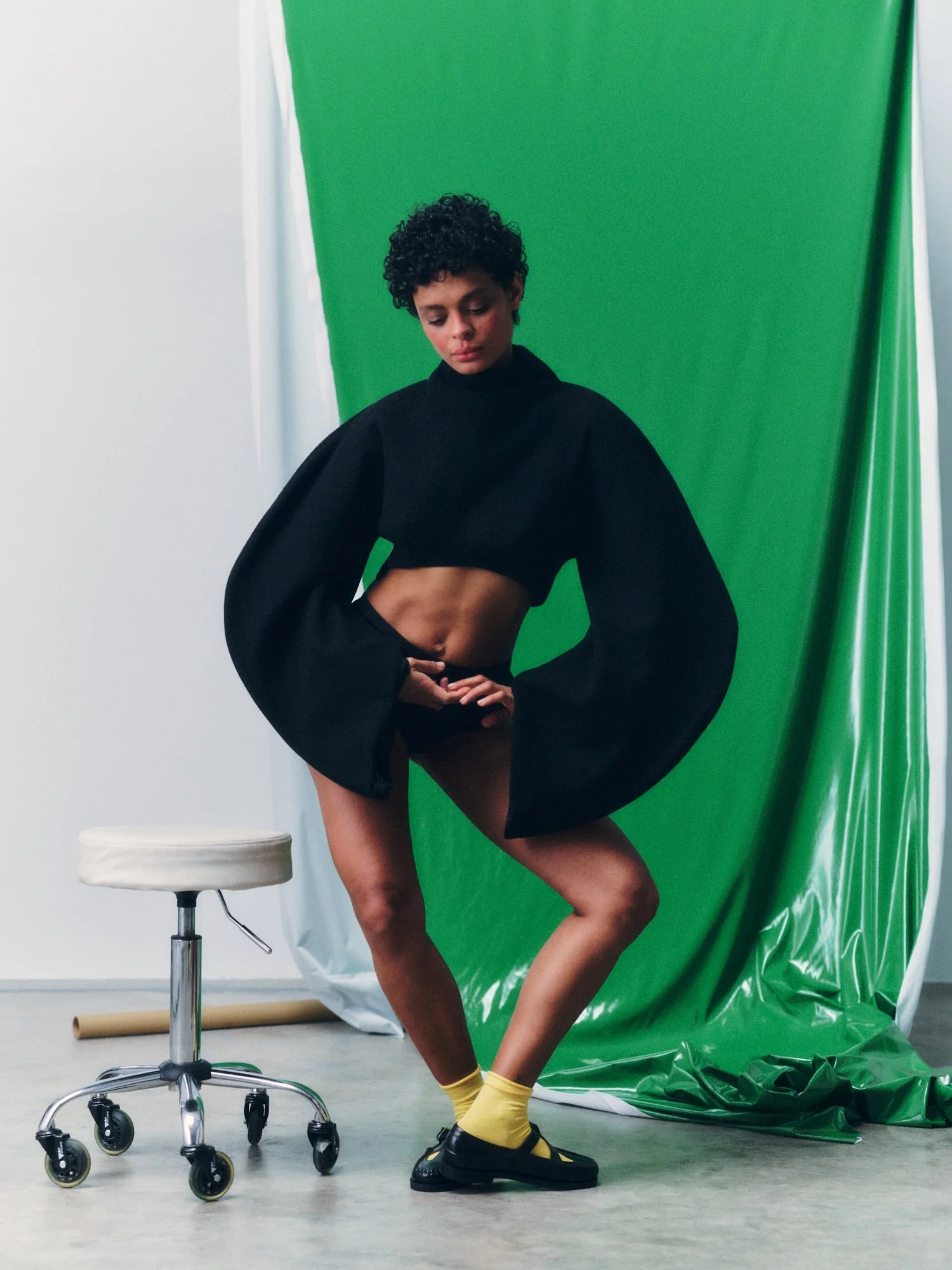 A woman with short curly hair and a black cropped hoodie standing with one leg raised, wearing yellow socks, black sandals, and standing next to a white rolling stool. Behind her is a green backdrop with white and green fabric on the sides.