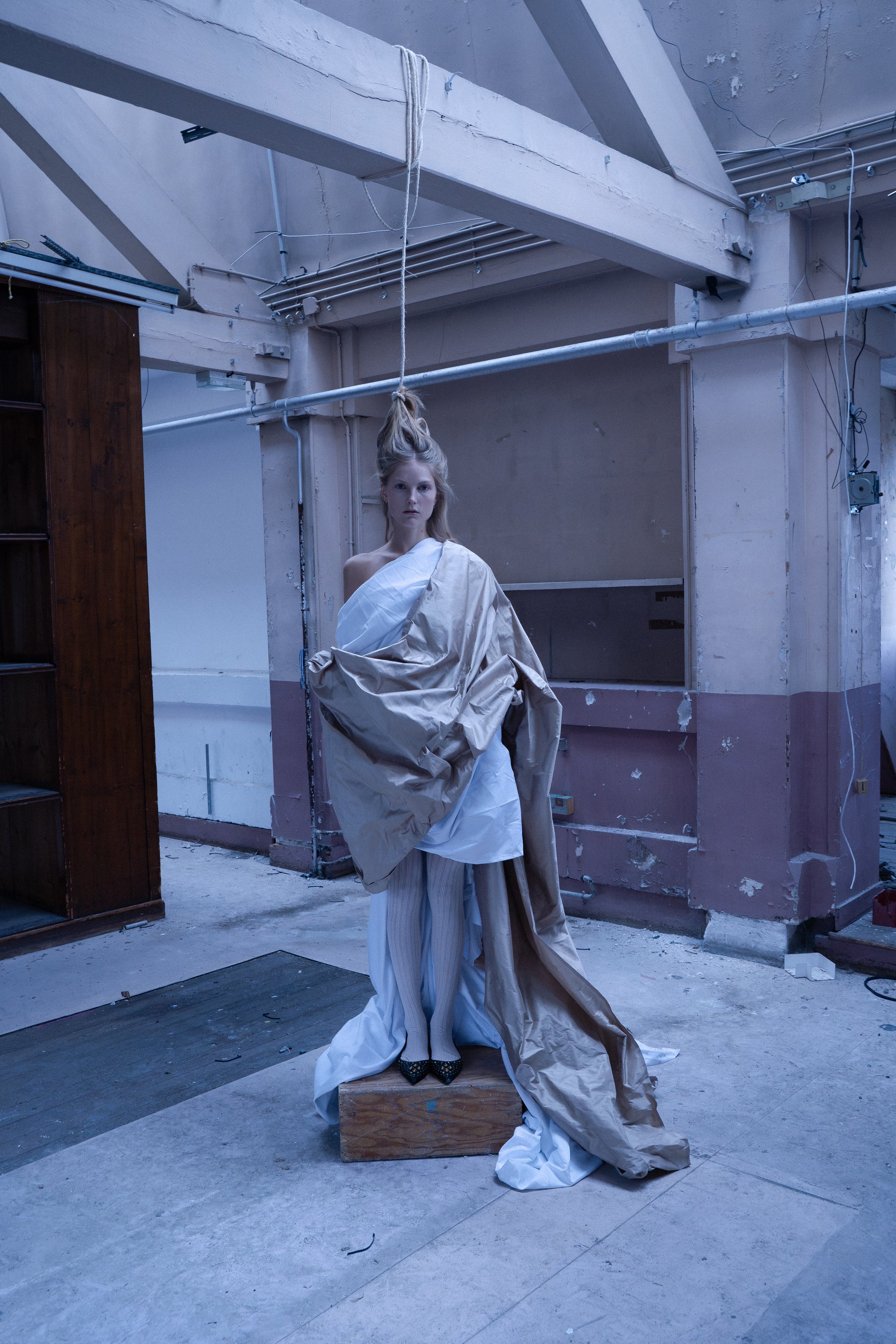 A fashion model stands on a wooden box in an industrial-looking room, wearing a layered outfit with draped fabrics and pinstripe pants, with an artistic hairstyle and minimal makeup.