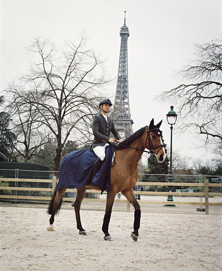 A person in equestrian attire riding a brown horse in an urban park with the Eiffel Tower in the background.
