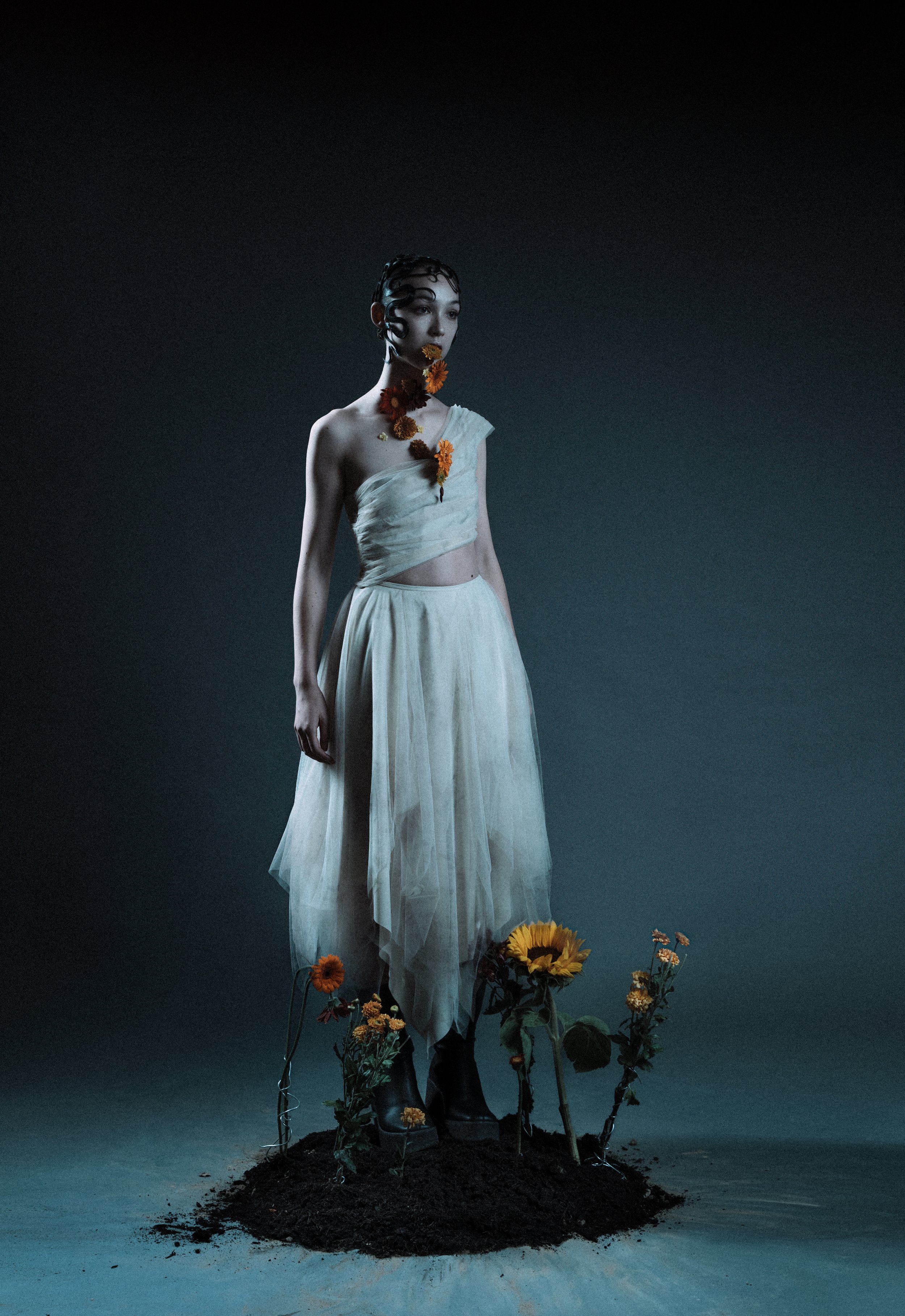 A woman wearing a white, flowing dress standing on a patch of soil surrounded by flowers, with dark background.