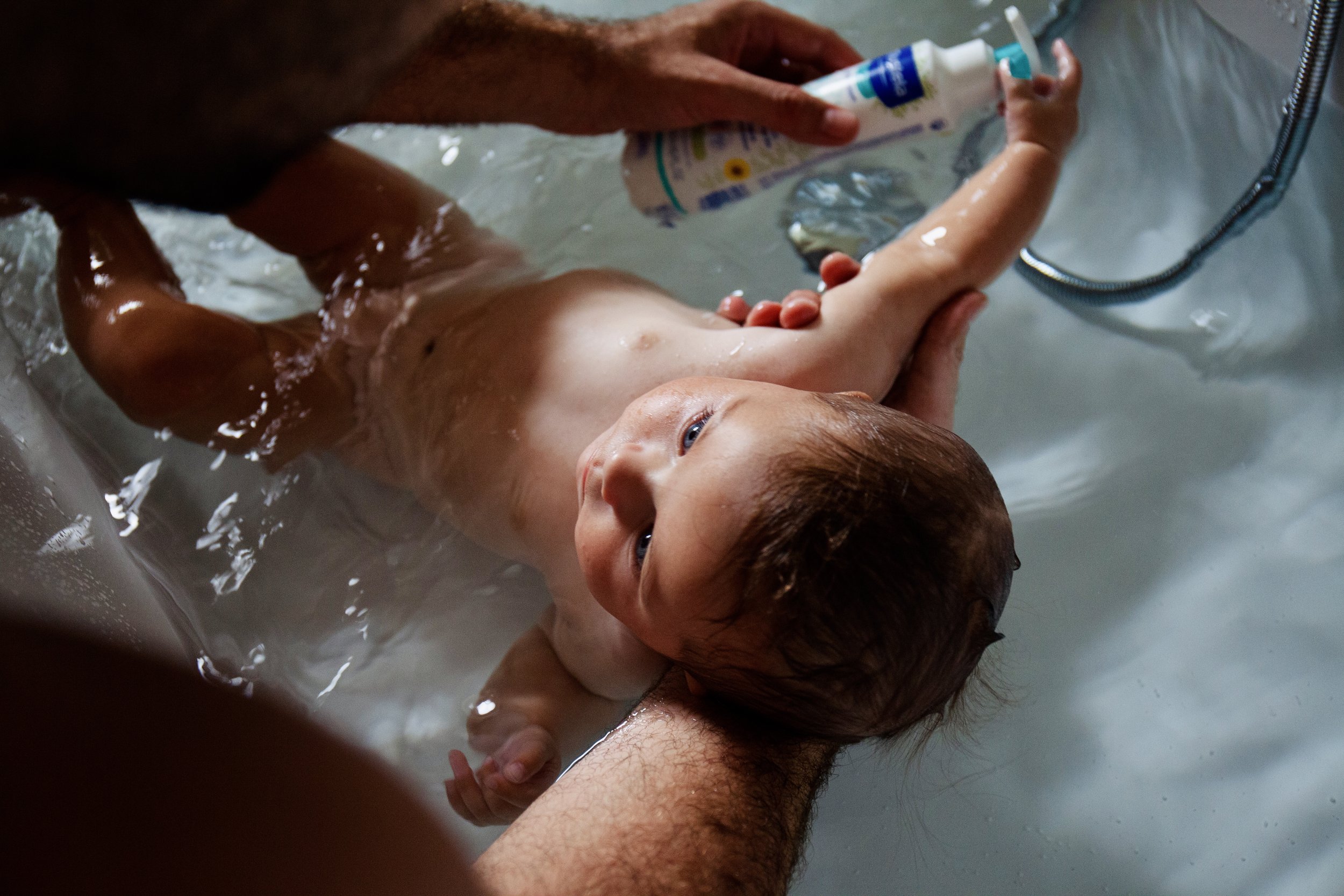 An adult giving a baby a bath in a sink or small tub with water. The adult is holding the baby steady while applying some sort of cream or lotion.