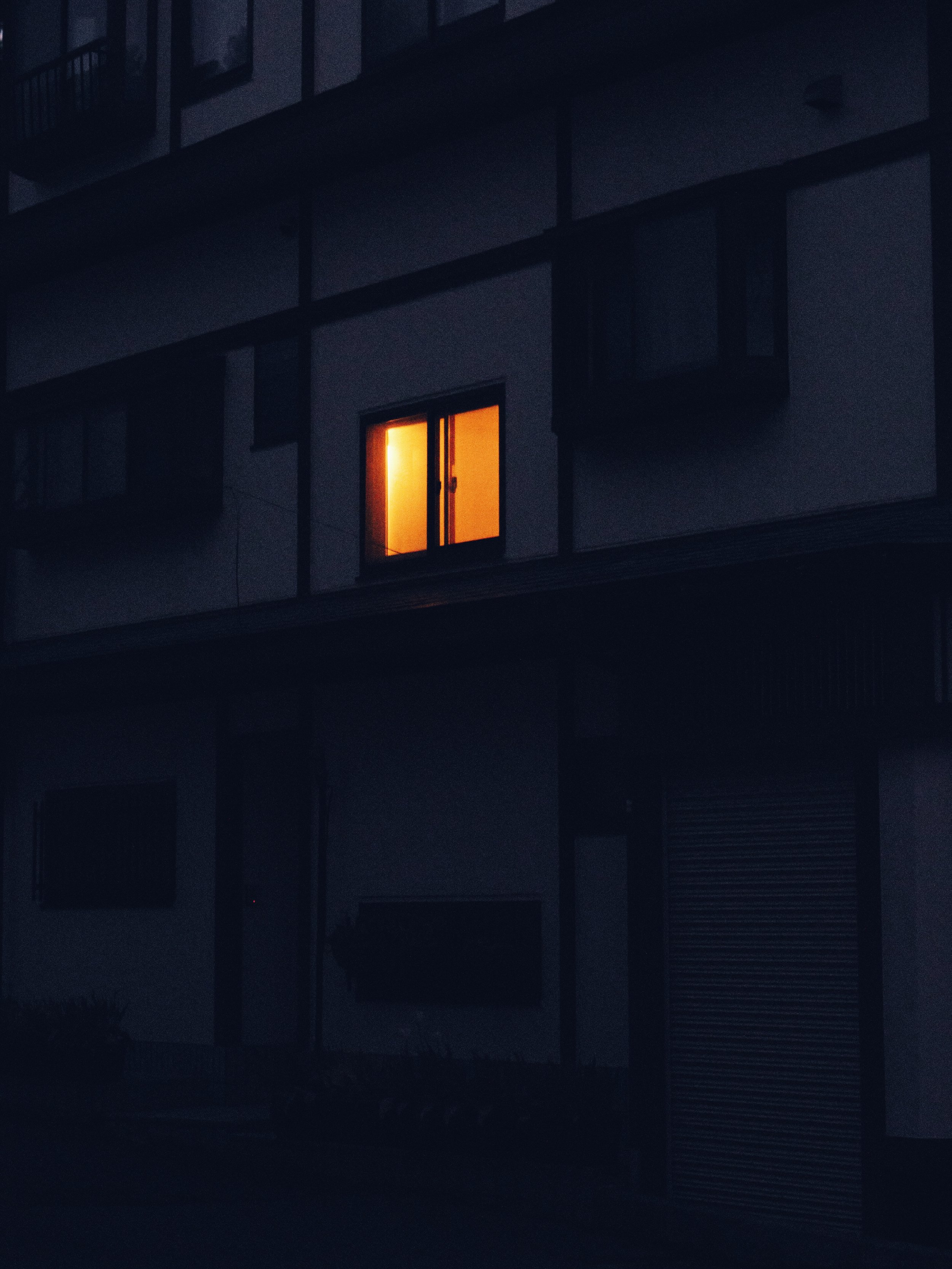 A dark exterior of an apartment building at night with a single illuminated window emitting a warm orange light.