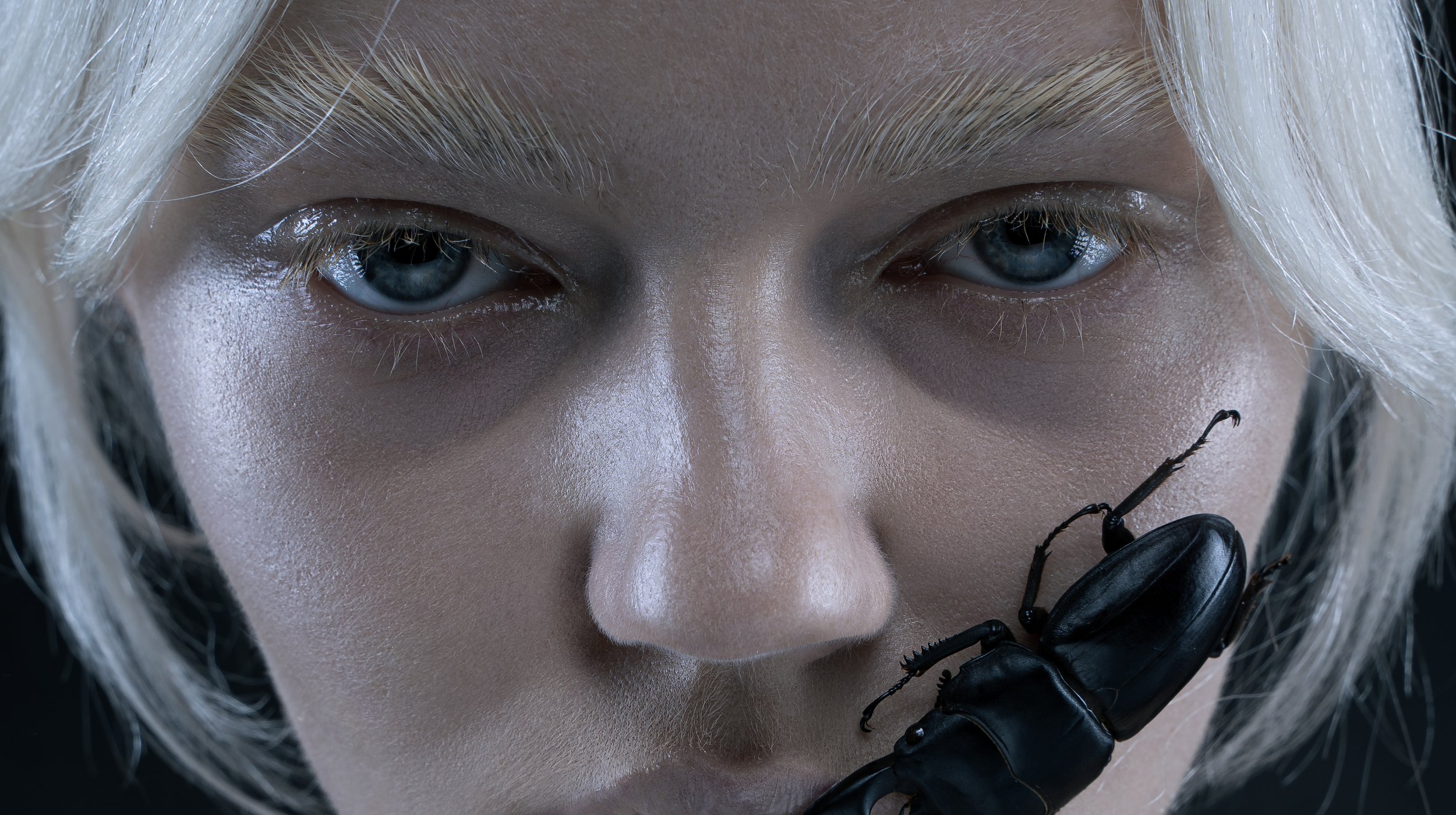 Close-up of a person's face with platinum blonde hair and blue eyes, holding a black beetle near their face.