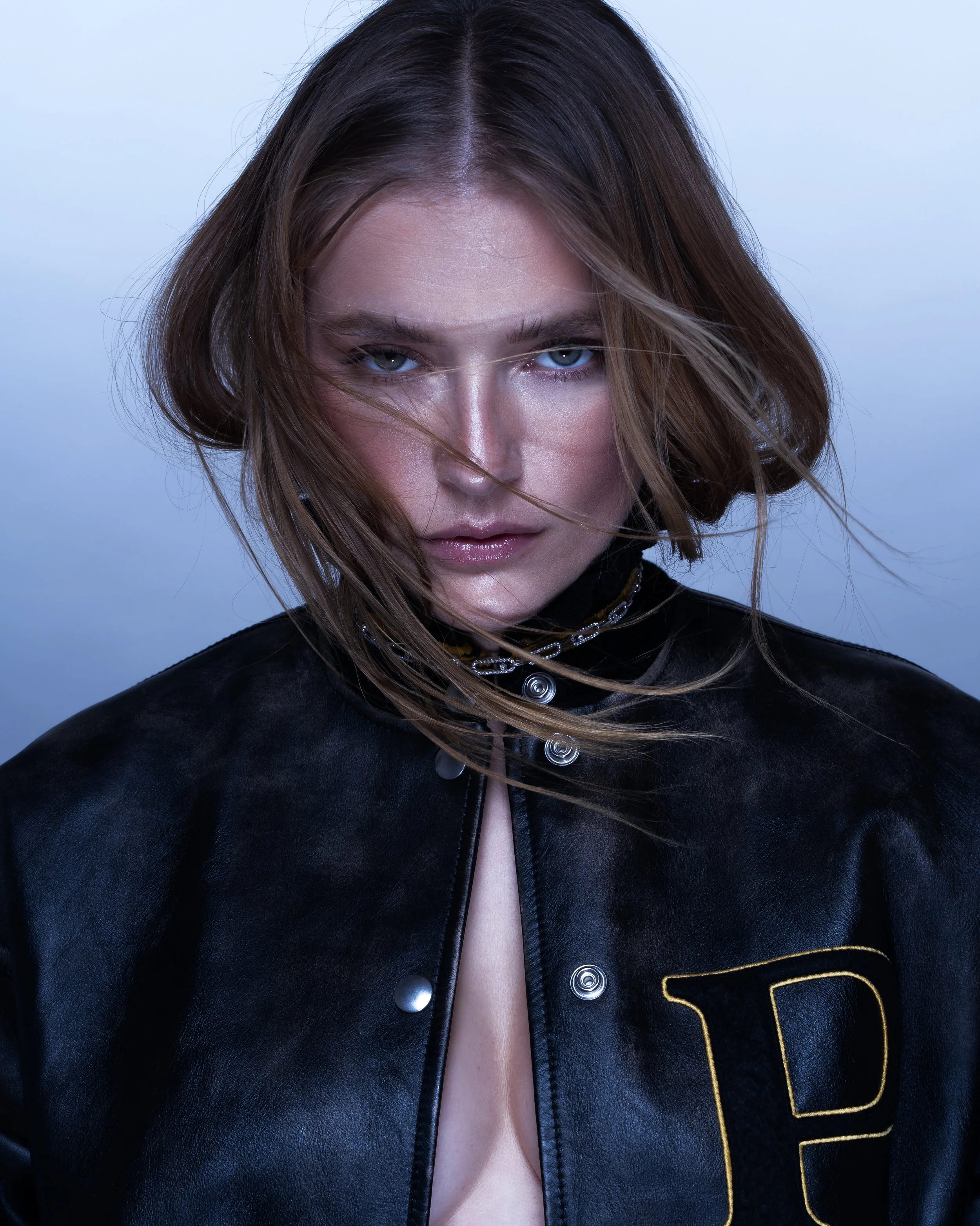 A woman with shoulder-length brown hair and blue eyes wears a black leather jacket with a large letter 'P' on it, and a chain necklace, looking directly at the camera