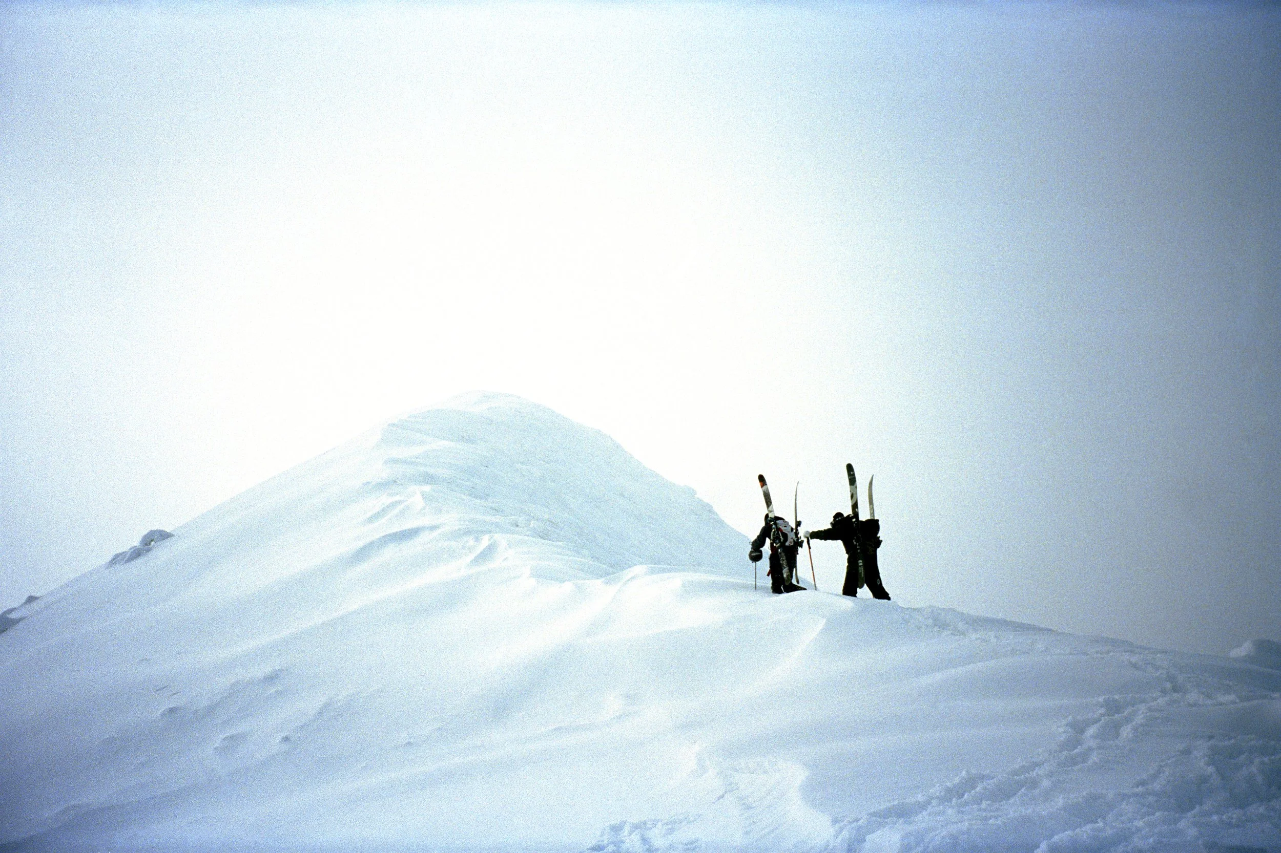 Two skiers hiking on a snow-covered mountain slope carrying skis on their shoulders.