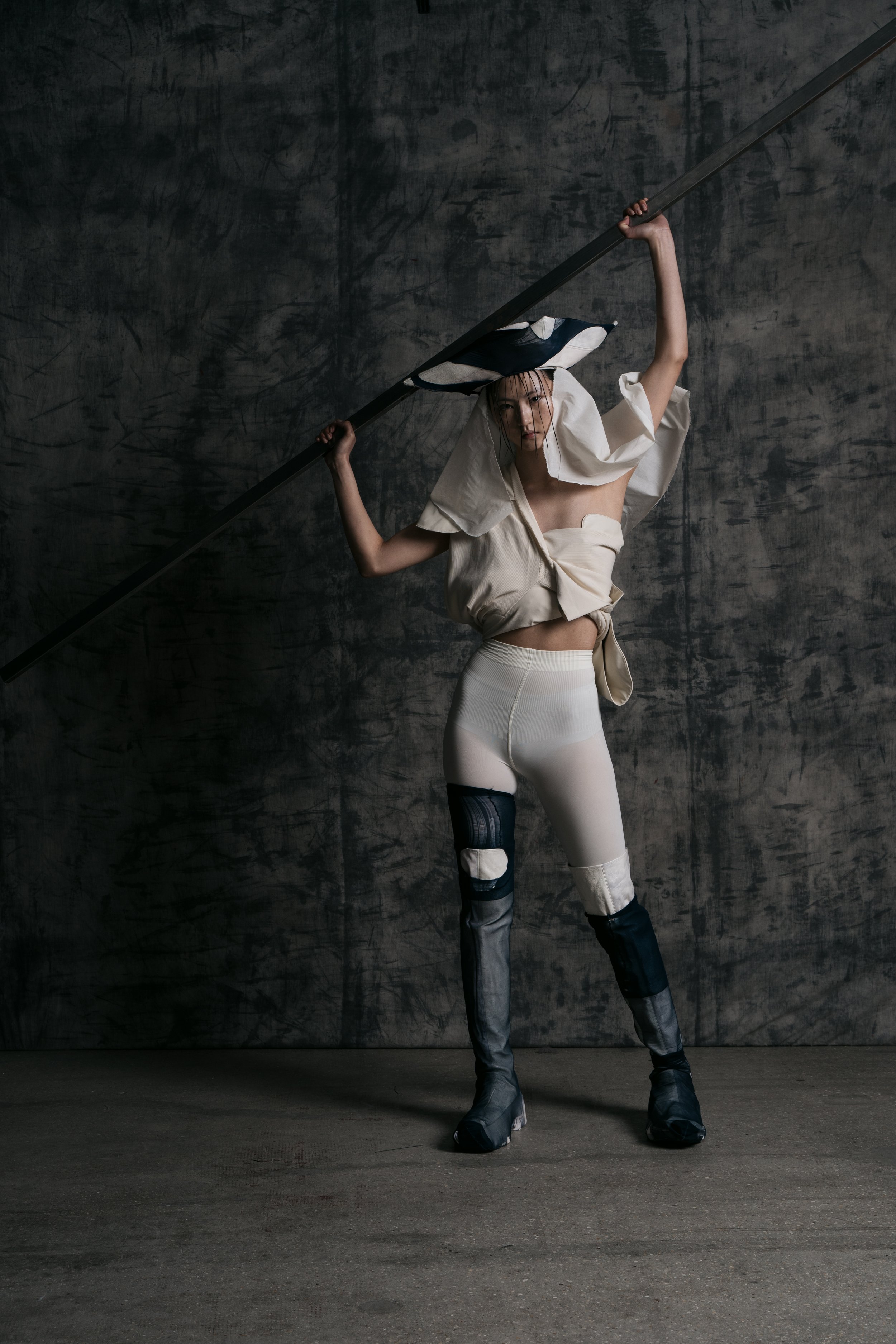 Fashion model with a spear, wearing an avant-garde outfit with a large hat and metallic pants, standing against a textured dark wall.