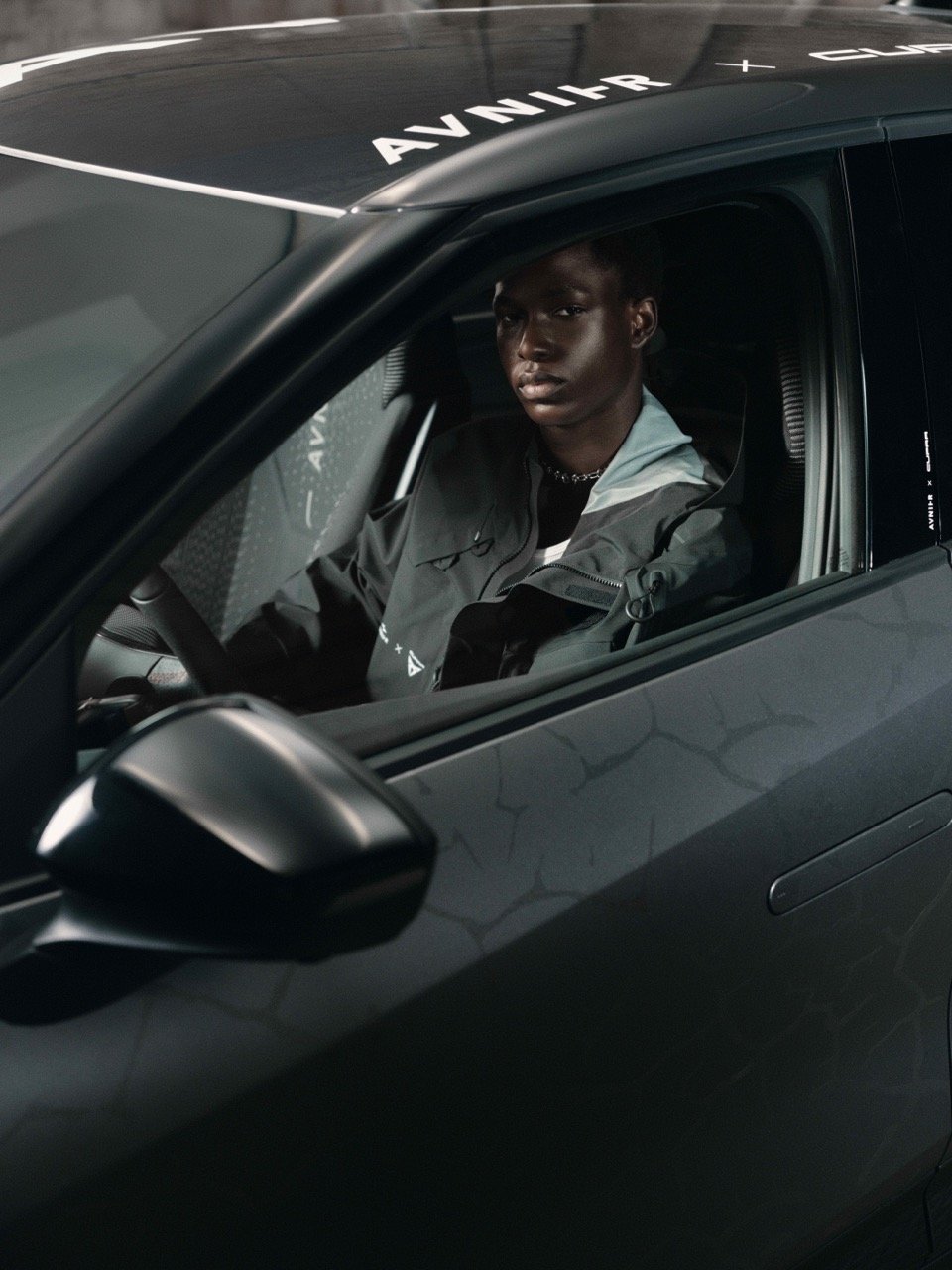 A young man sitting inside a black sports car with 'AVENIR' written on the roof, looking out the window.