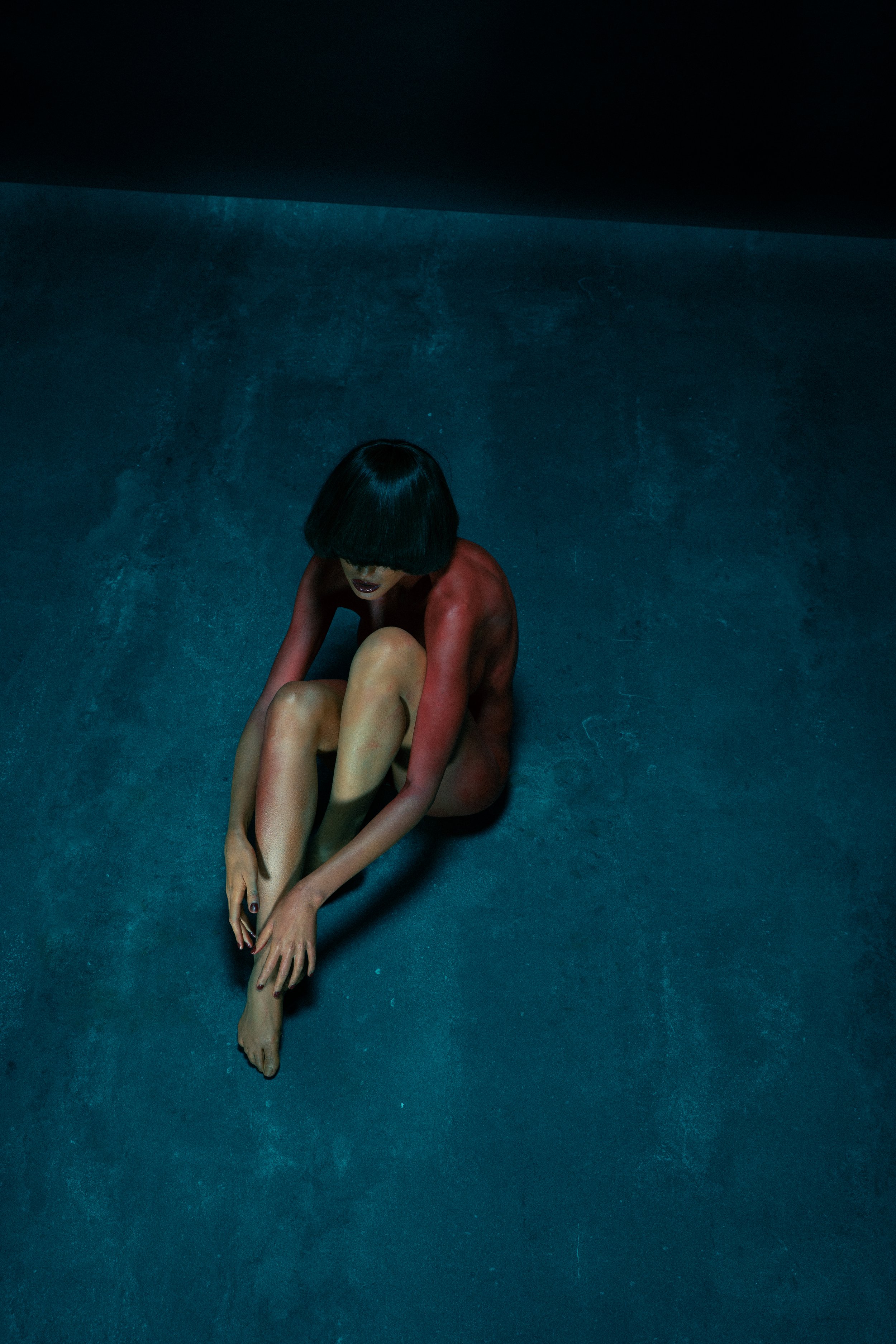 A woman with short dark hair sitting on a dark textured floor, with her knees pulled towards her chest, in a dimly lit room with dark walls.
