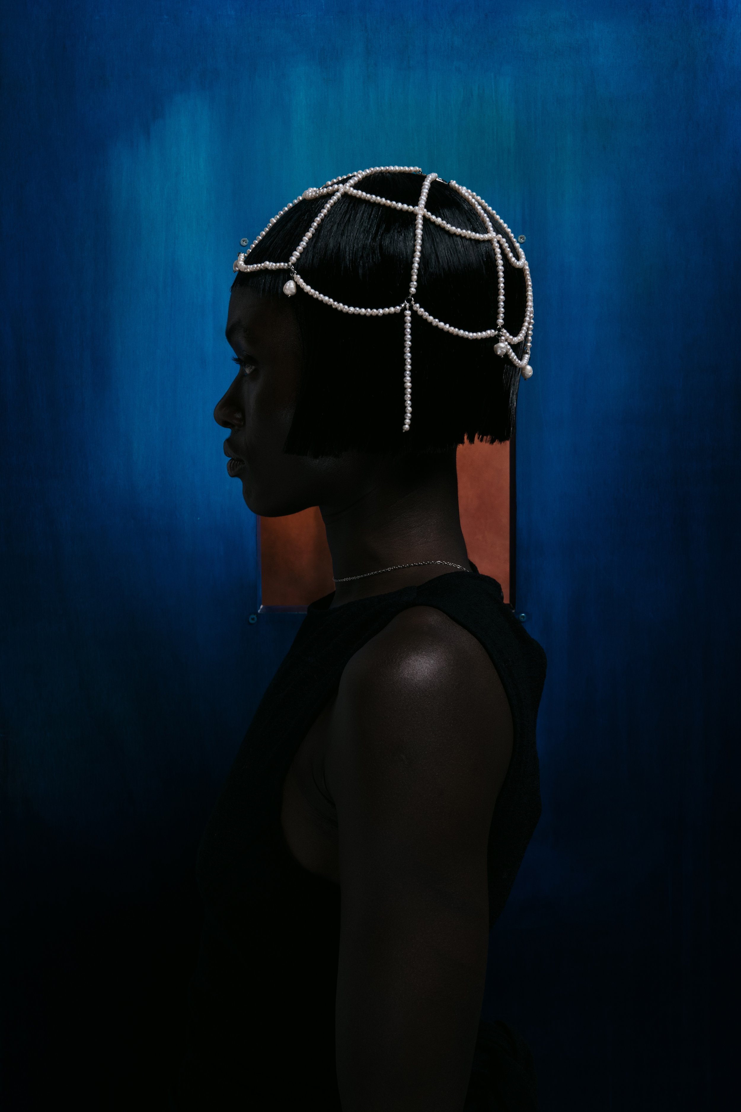 A woman with short black hair and dark skin, wearing a pearl headpiece and a black sleeveless top, standing in profile against a colorful abstract background.