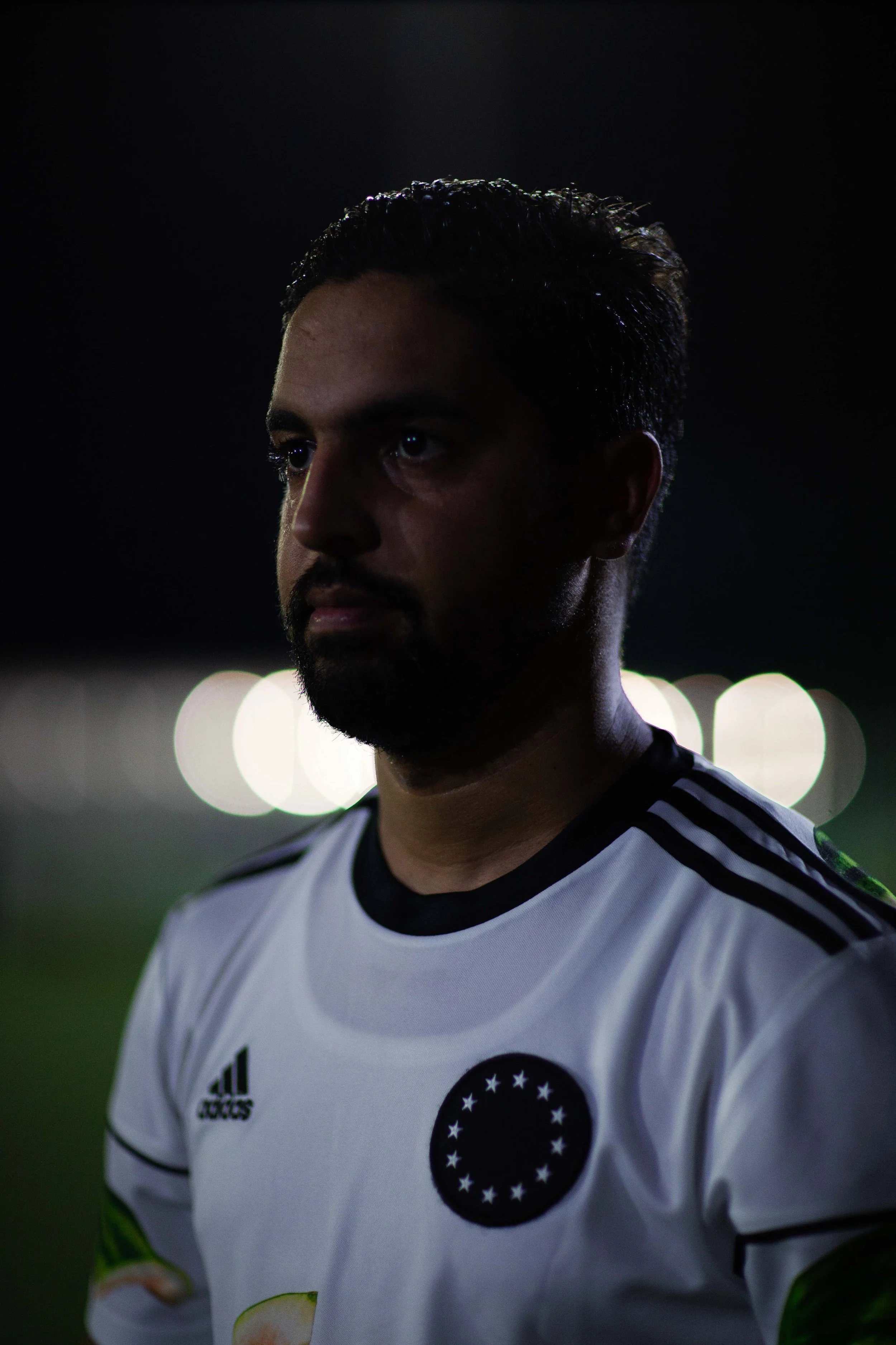 A man with dark hair, beard, and mustache wearing a white sports jersey with black accents and a circular logo with stars, standing outdoors at night.