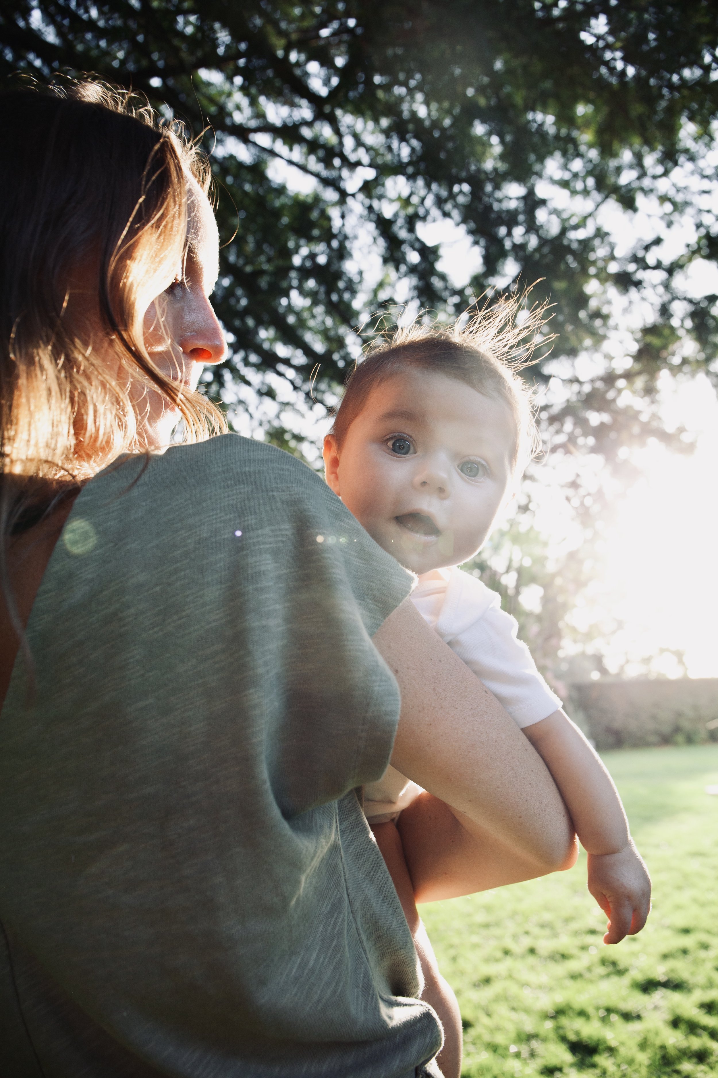 A woman holding a young child outdoors in a park, with sunlight filtering through trees in the background.