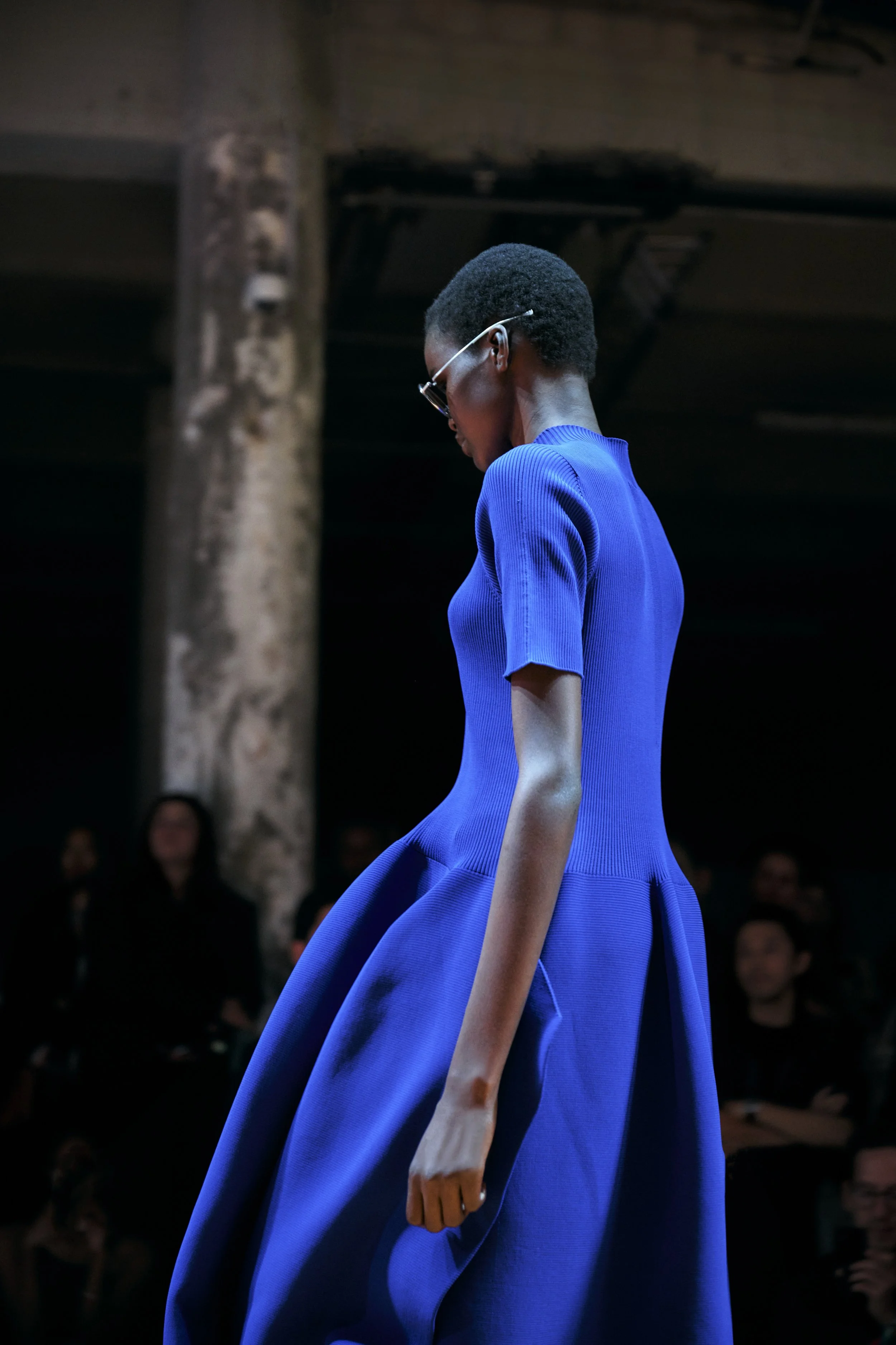 A fashion model in a bright blue dress walks on a runway during a fashion show, with an audience watching in the background.