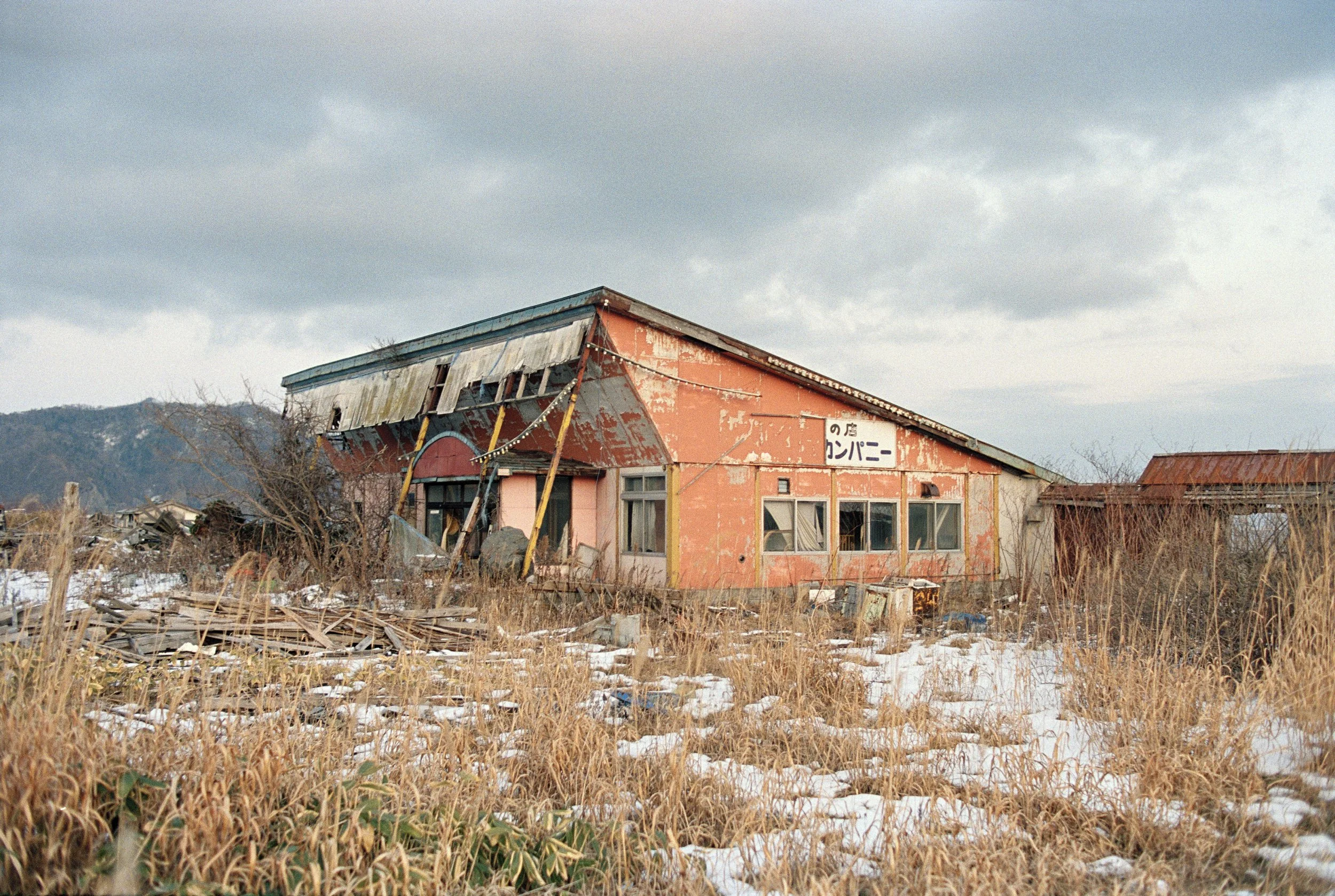 An abandoned, dilapidated building with peeling orange paint, broken windows, and a sagging roof, surrounded by overgrown grass and debris, with a cloudy sky overhead.