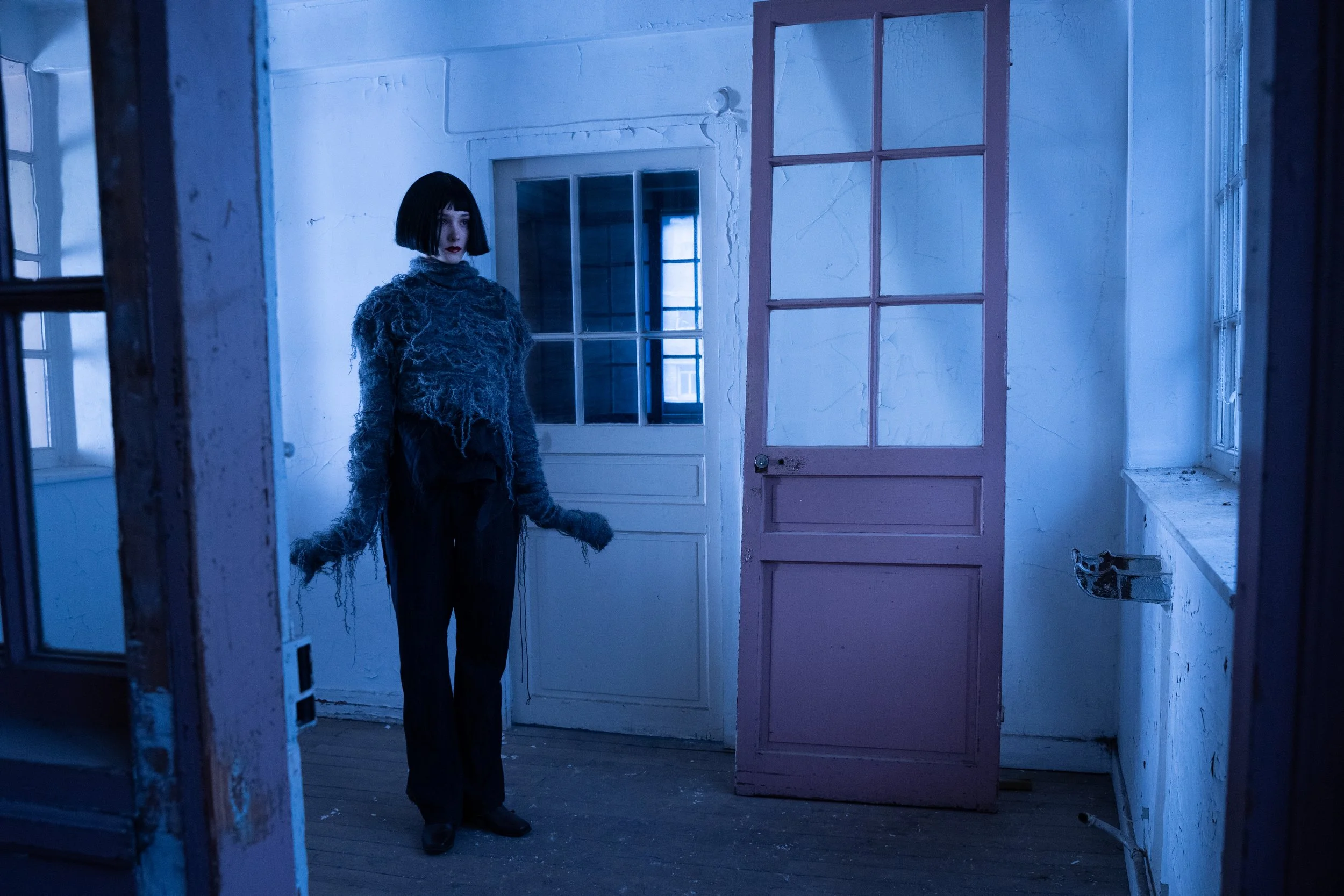 A woman with short black hair and dark makeup standing in a dimly lit, abandoned room with peeling walls and worn wooden floor. She wears a fuzzy, gray, ripped sweater and black pants, looking towards the camera with an intense expression. The room h