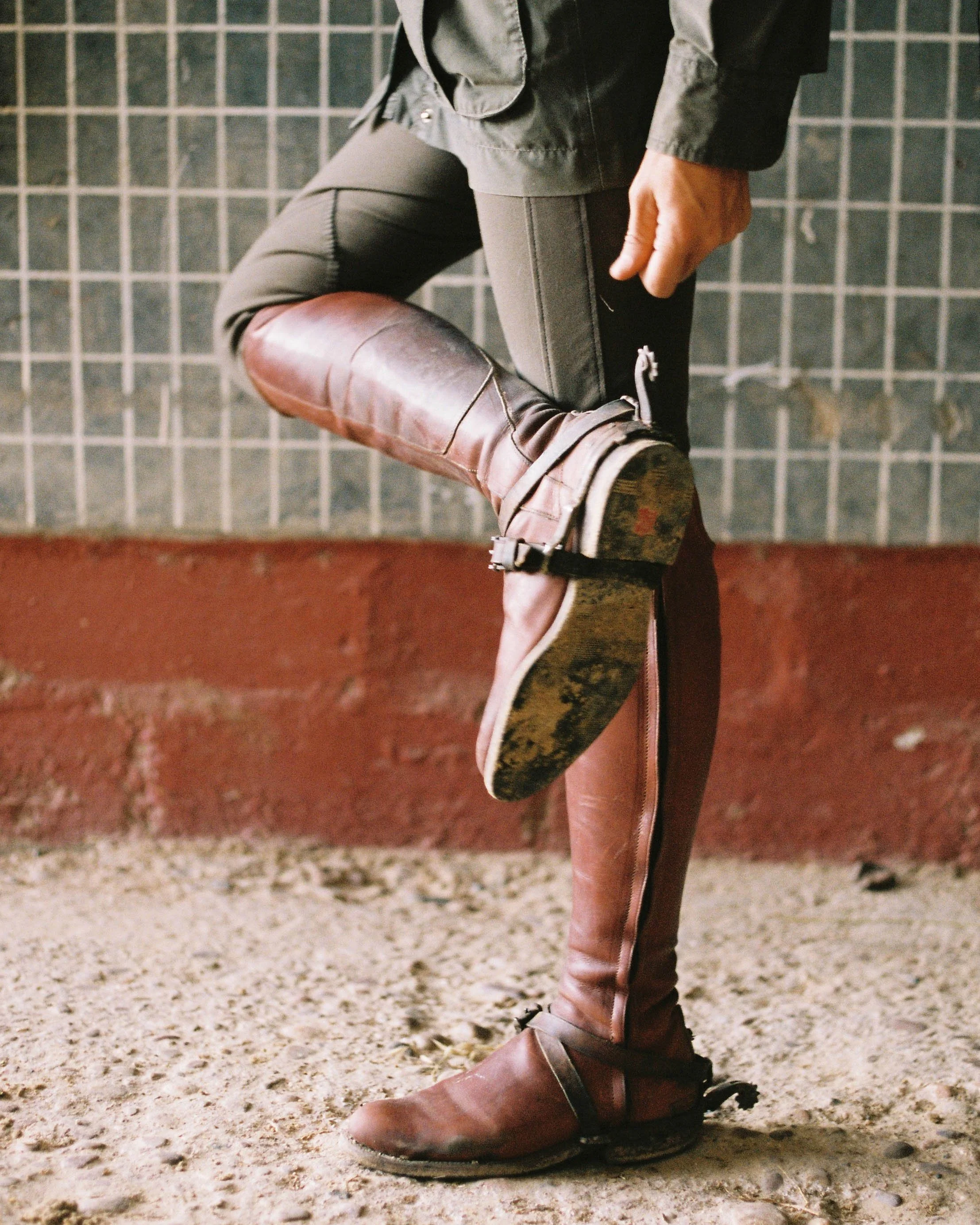 Person wearing brown cowboy boots and dark pants, standing on a gravel ground against a tiled wall background.