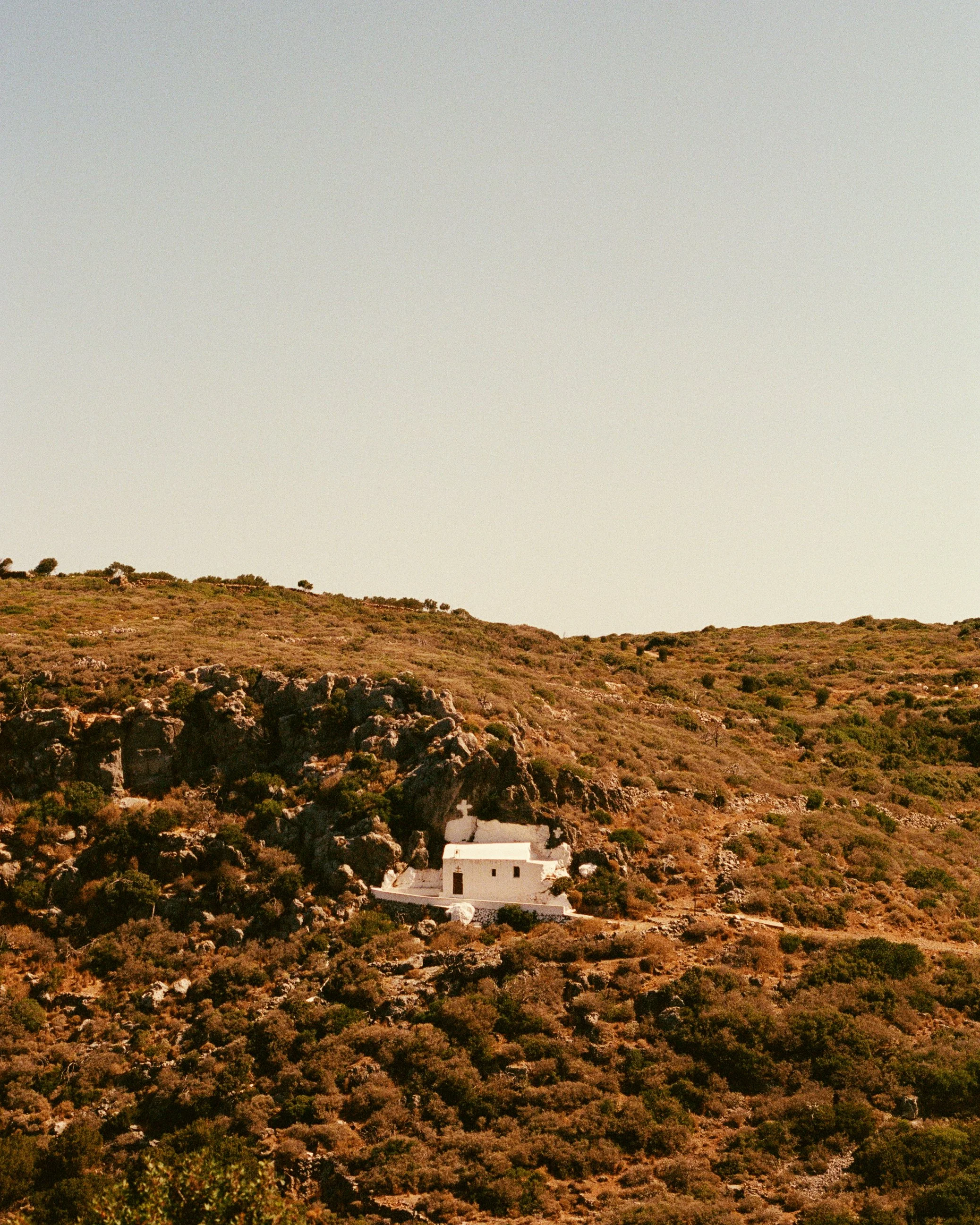 Small white church with a cross on the roof, situated on a hillside with sparse vegetation and rocks, under a clear sky.