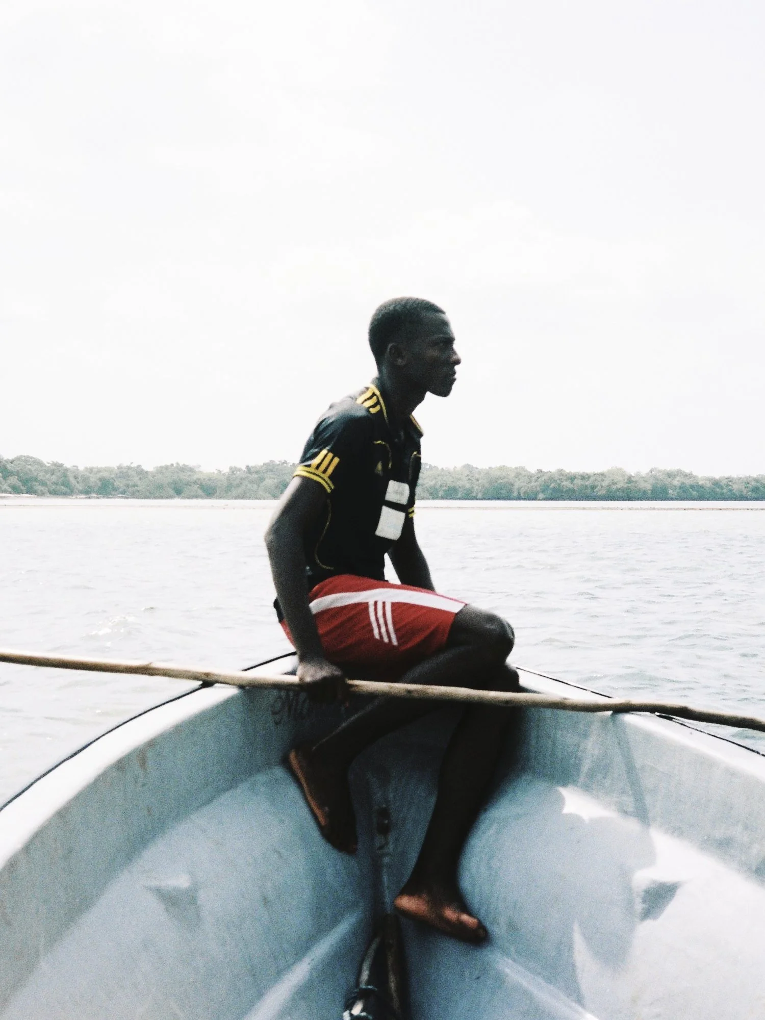 A young man wearing a black jersey with yellow accents and red shorts sitting on the edge of a boat, holding an oar, on a body of water with land and trees in the background.