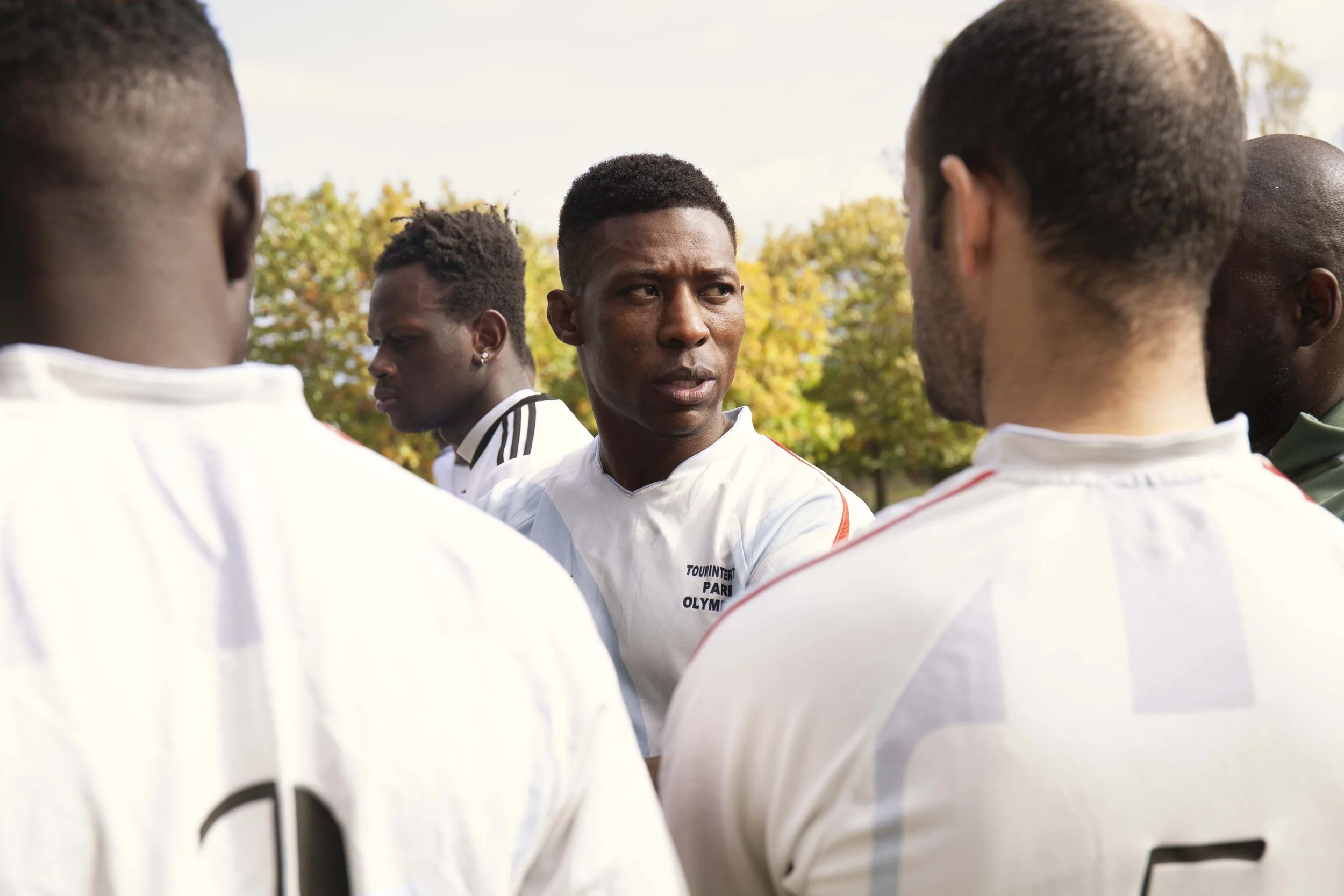 A group of diverse soccer players in white jerseys gathered around a coach or teammate, who is speaking to them, outdoors on a field with trees in the background.