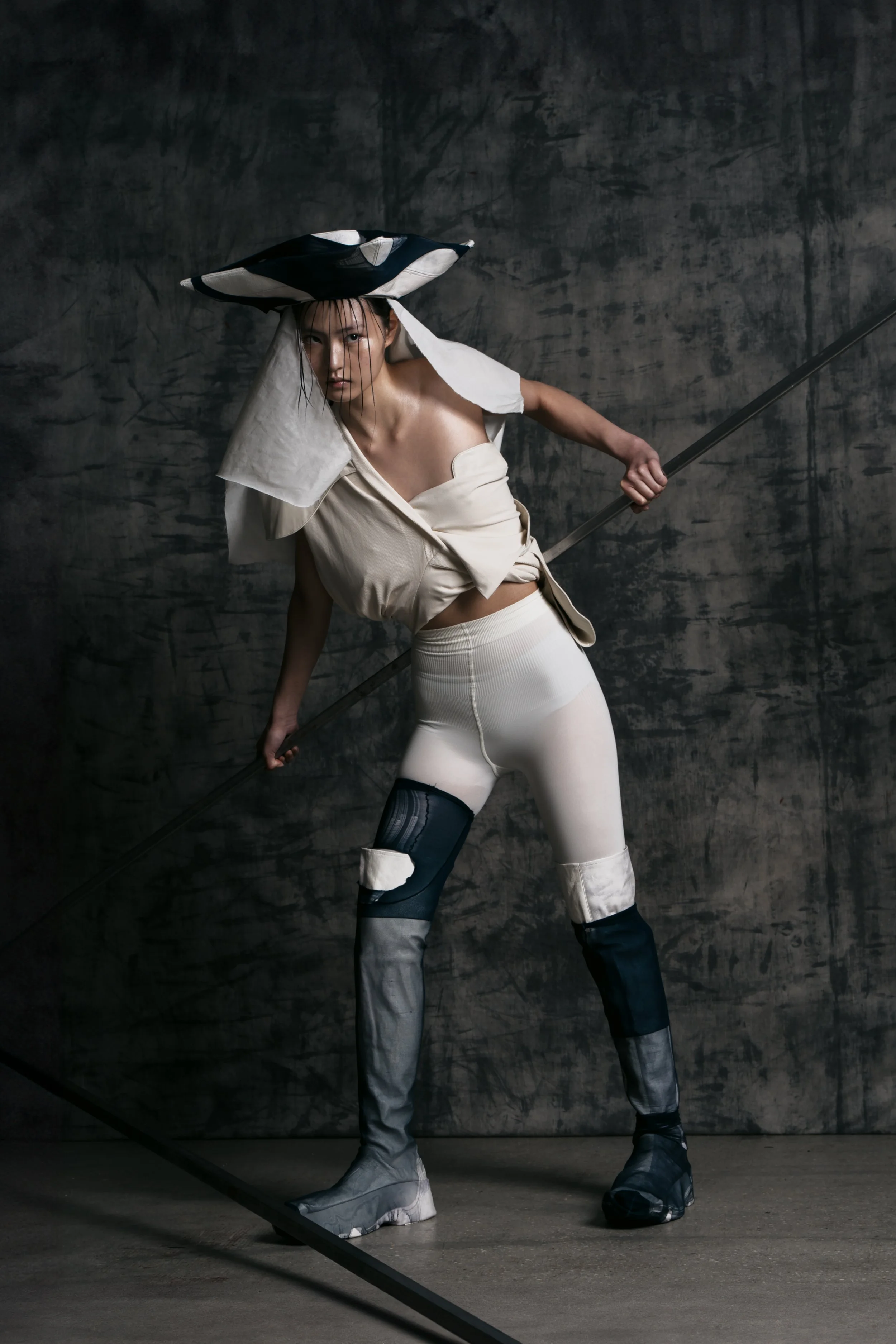 A woman dressed in fashion-forward clothing, holding a long stick or pole, standing in front of a dark, textured wall.