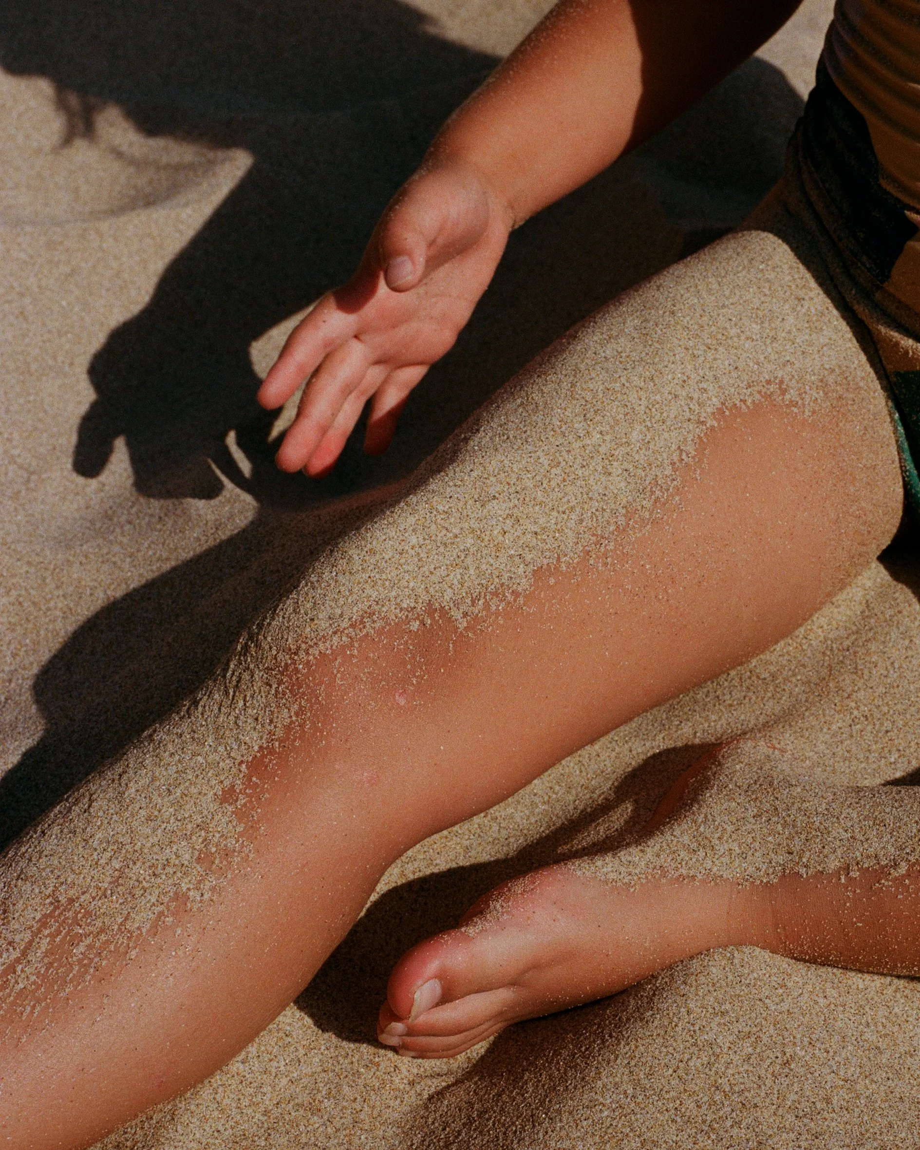 A person with sand on their leg and hand, sitting on a sandy surface, with a shadow cast on the sand.
