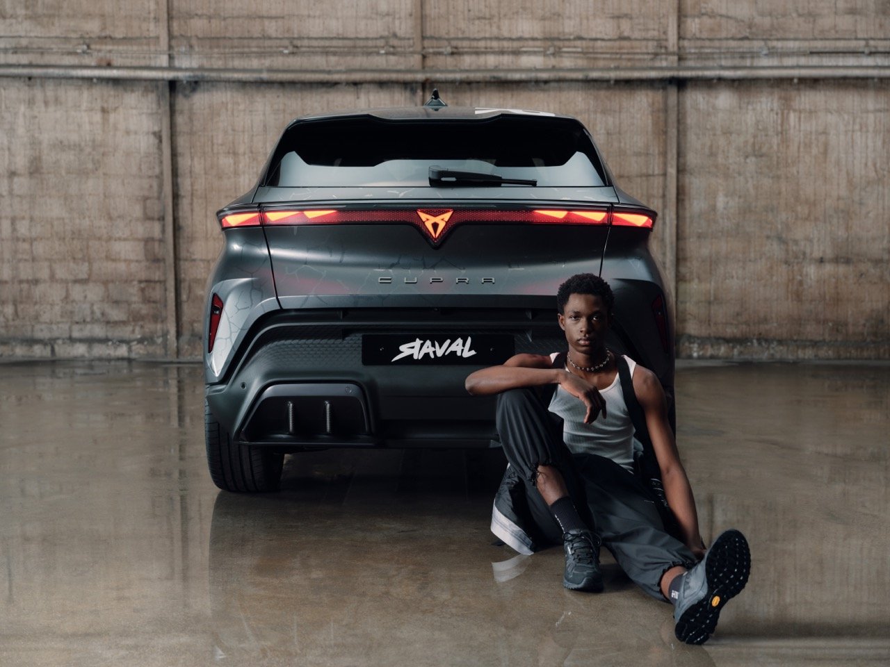 A young person with short curly hair, wearing a white tank top and black pants, sitting on the ground with one leg bent and the other extended, in front of a black luxury SUV with an illuminated logo and sleek design, in an industrial space with conc