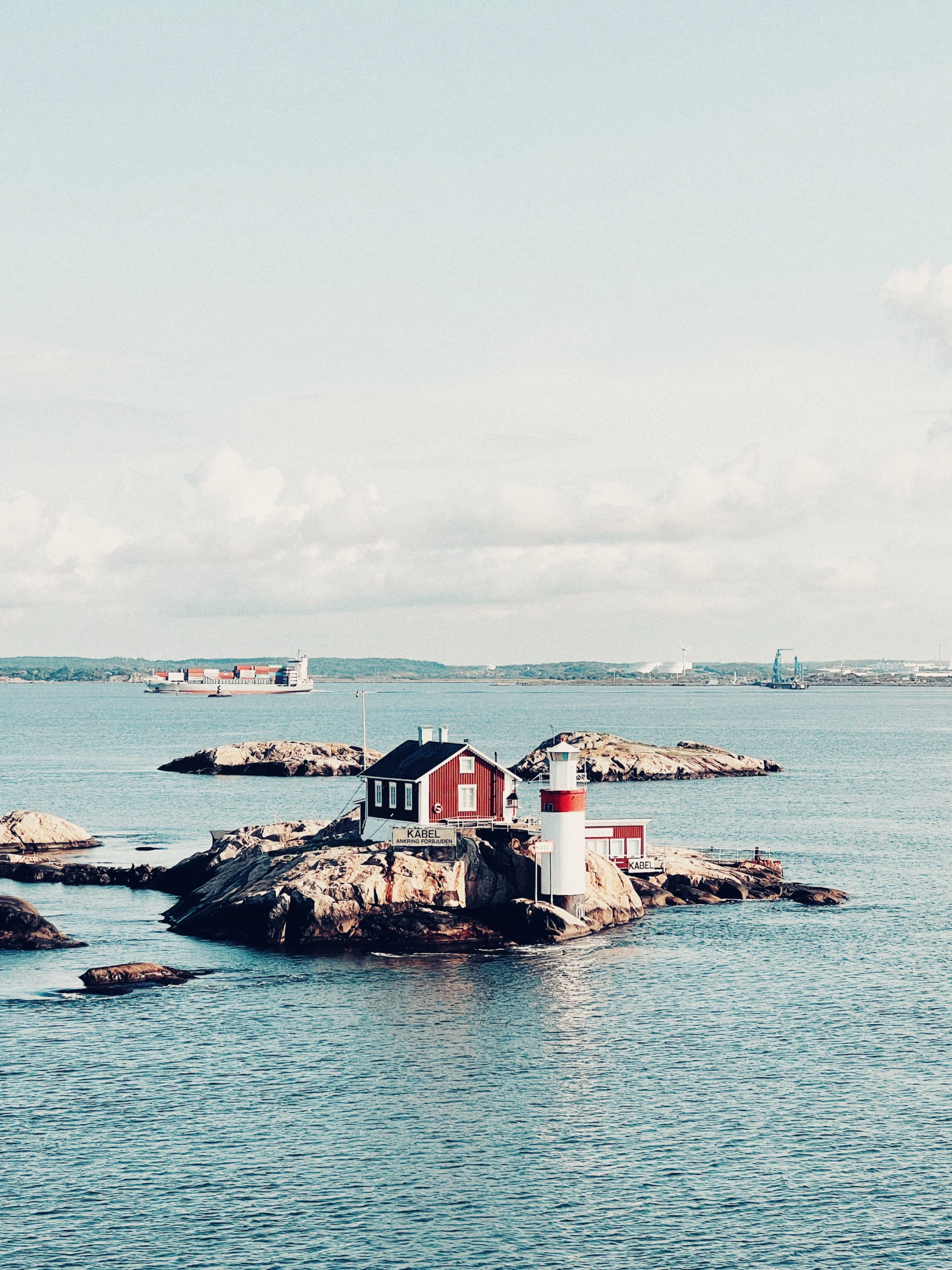 A small red and white lighthouse on a rocky island surrounded by calm water, with a house and signs that say "KABEL".