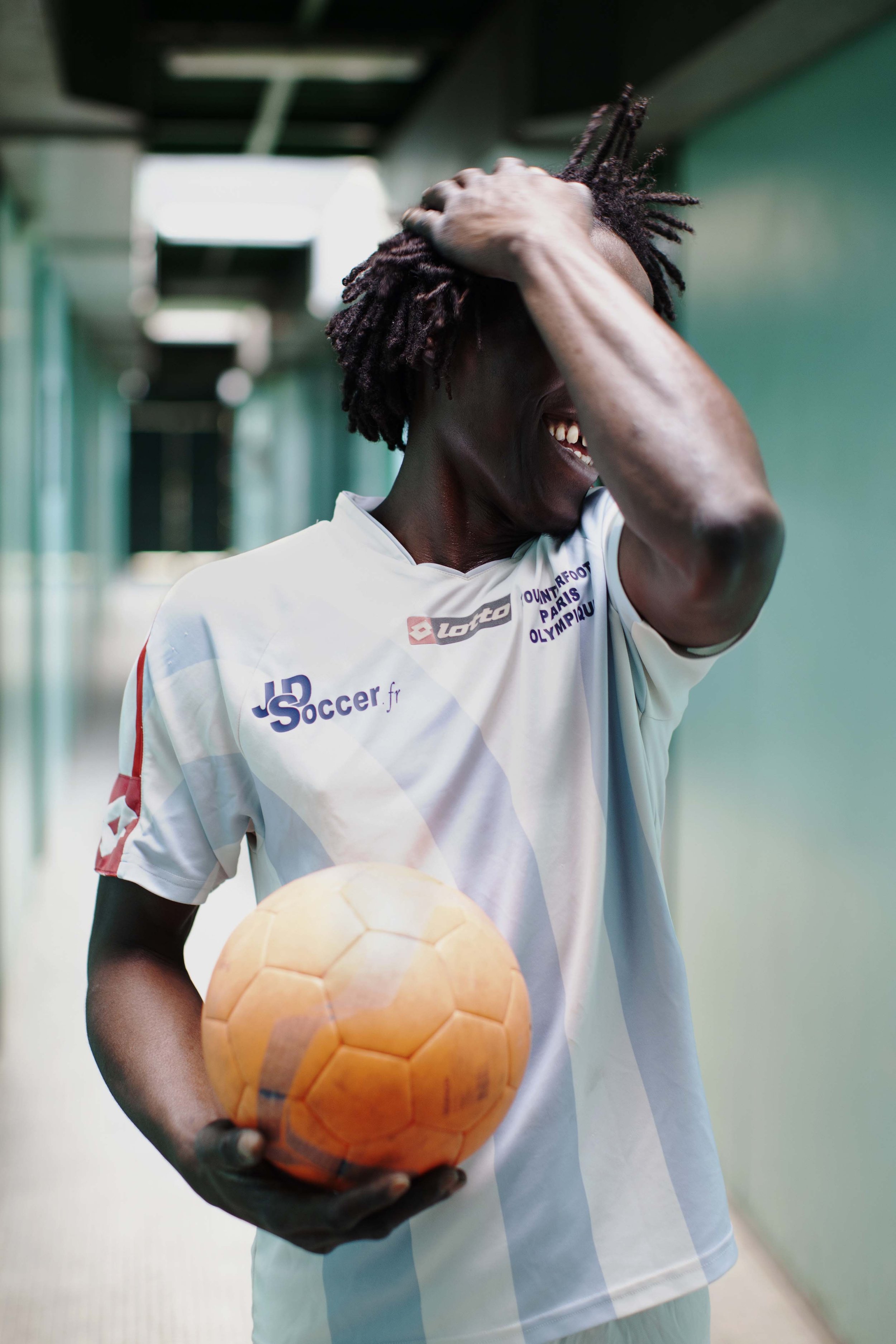 A man with dreadlocks wearing a white soccer jersey laughing, holding an orange soccer ball, in a hallway.