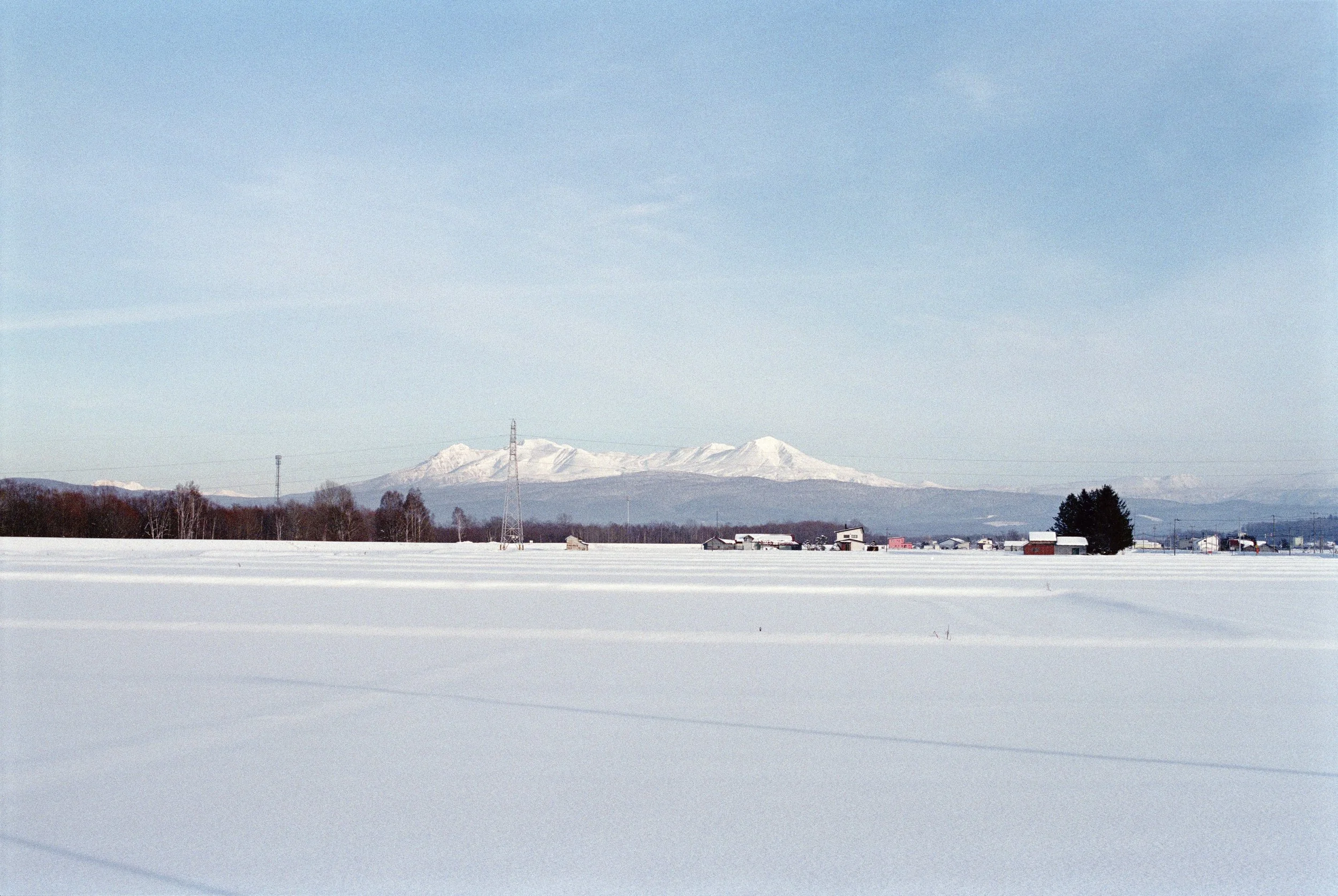 Snow-covered field with a few houses and a snow-capped mountain in the background under a blue sky.