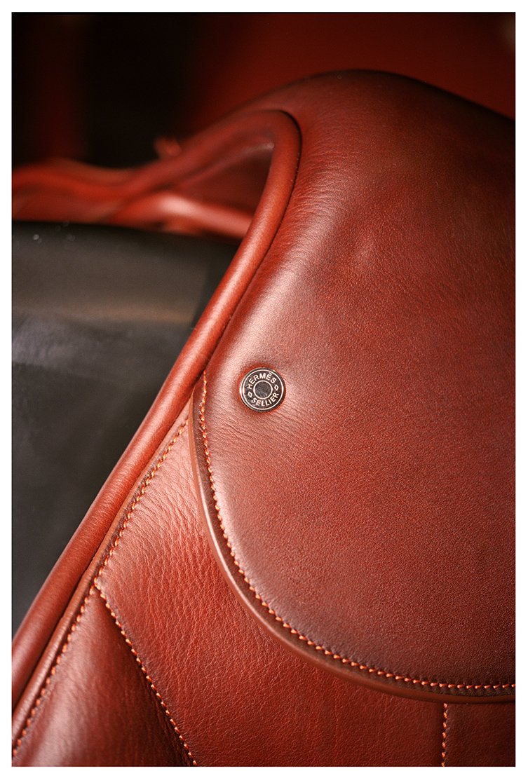 A close-up of a Hermès saddle leather handbag in reddish-brown color, showcasing detailed stitching and a metal badge with the Hermès and Sadelier branding.