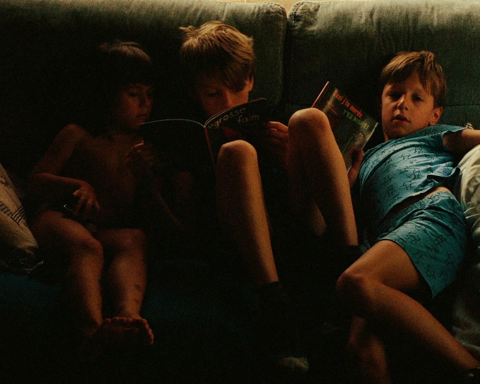 Four children sitting on a dark couch, reading and relaxing in a dimly lit room.