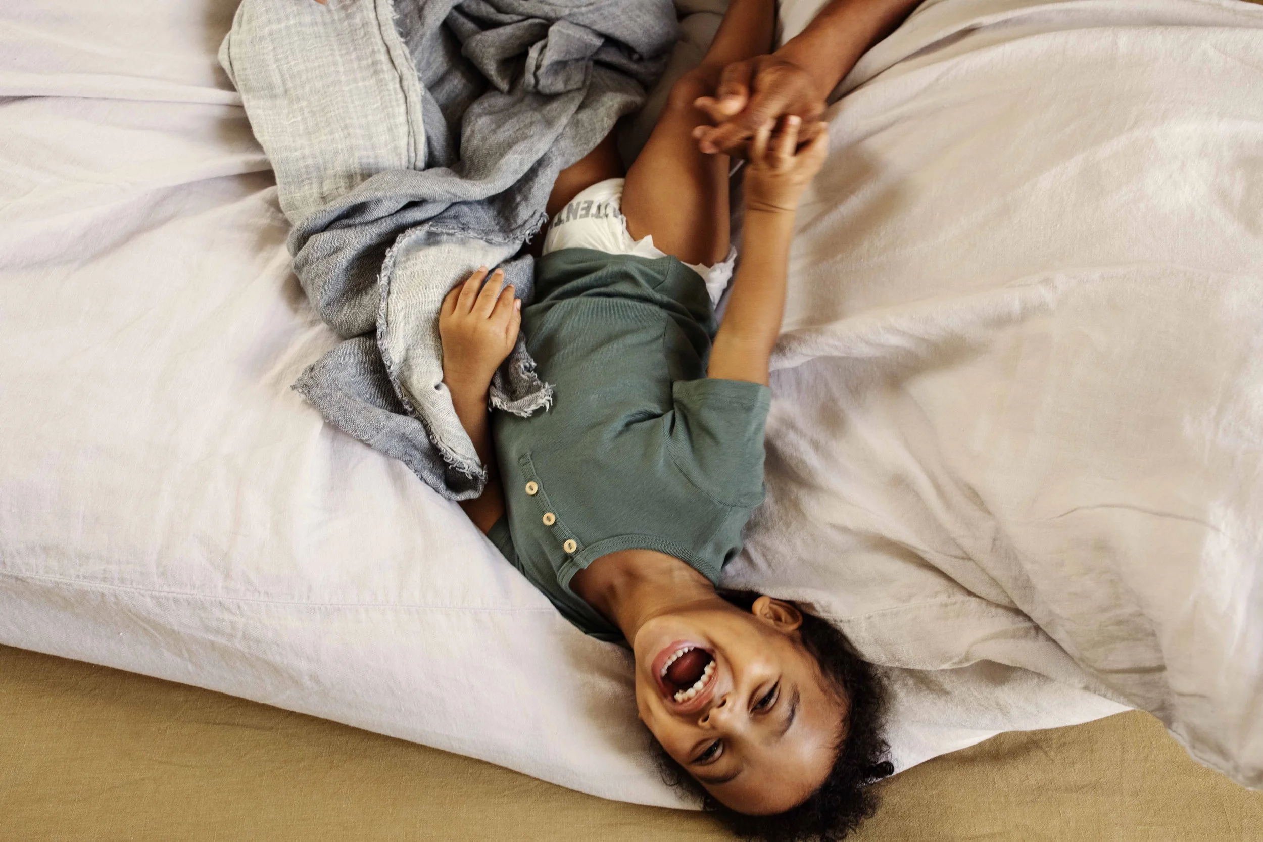 A young girl lying on a bed, laughing with her mouth open, holding hands with an adult beside her.