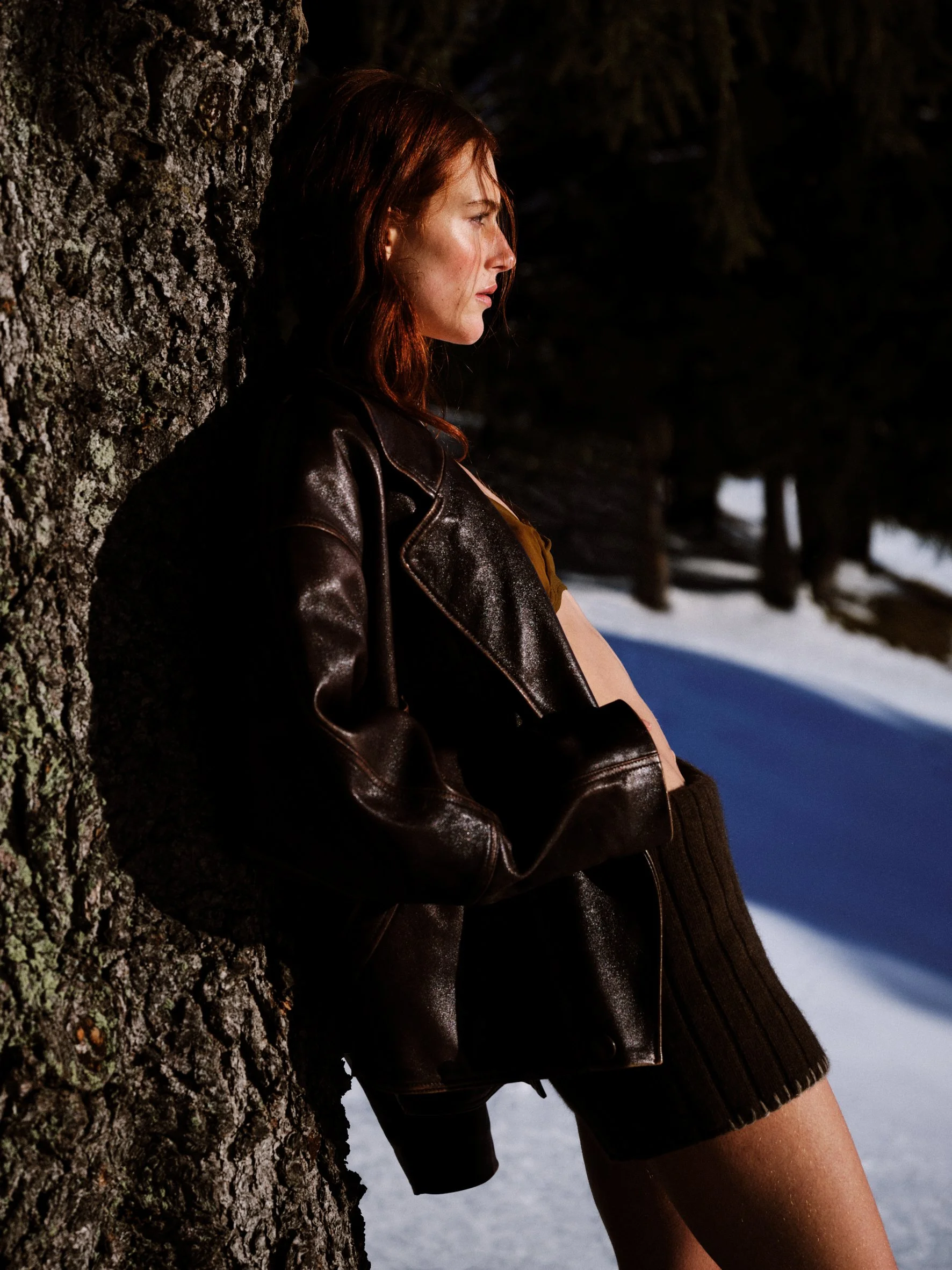 A woman with red hair leaning against a tree in a snowy outdoor setting, wearing a leather jacket and a skirt.