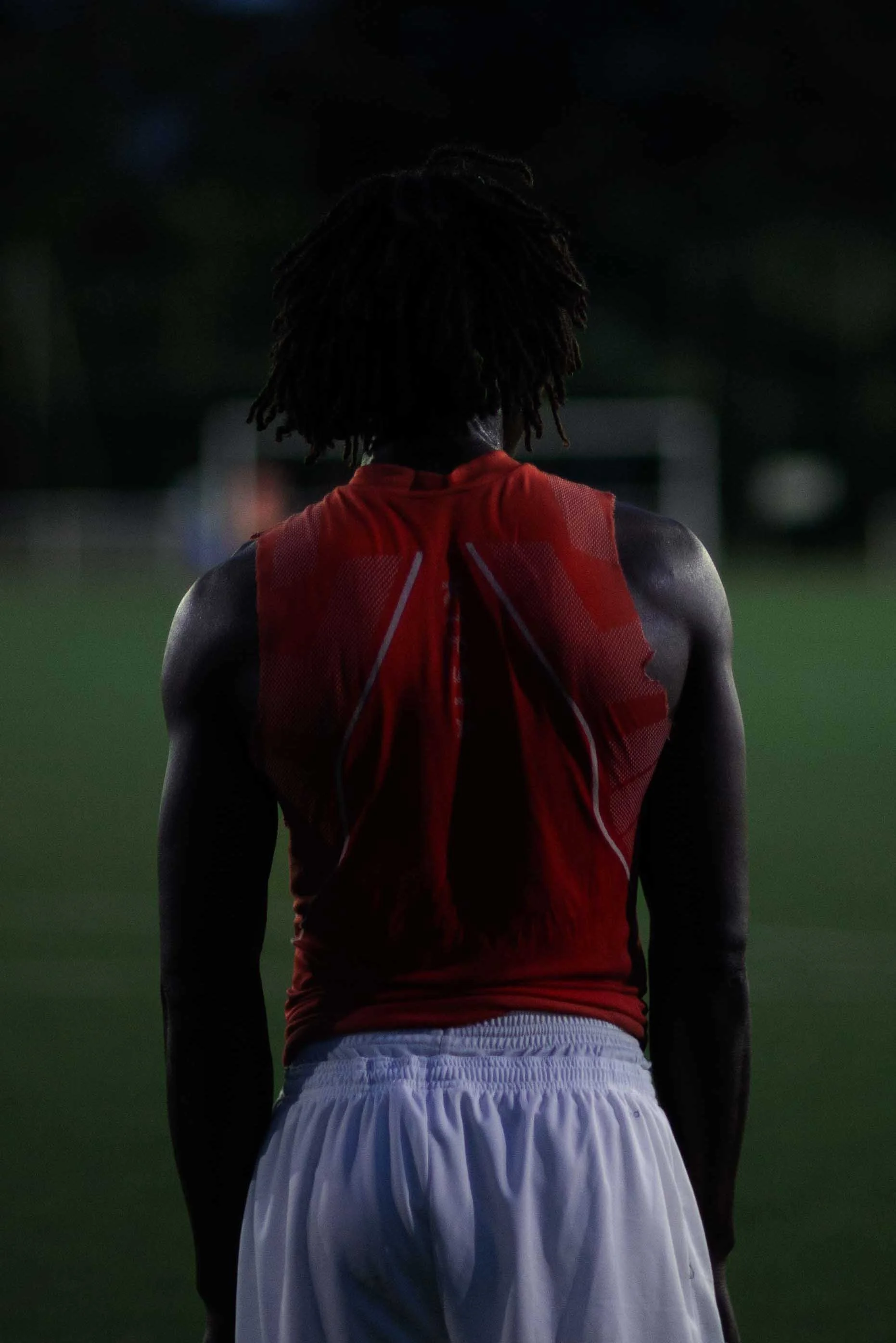 Athlete with dreadlocks wearing a red sports jersey and white shorts, seen from the back, standing on a field at dusk or nighttime.