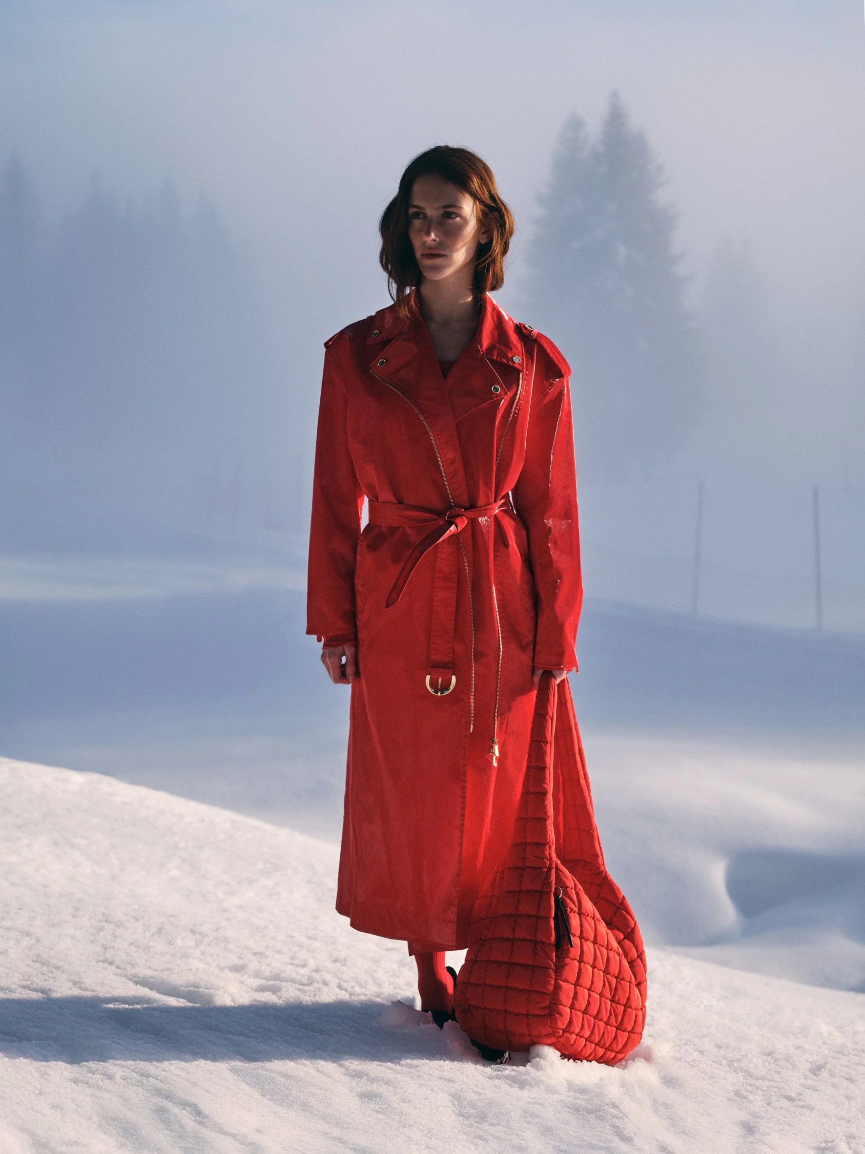 A woman with shoulder-length auburn hair standing in snowy landscape wearing a long red coat, holding a matching red quilted bag.