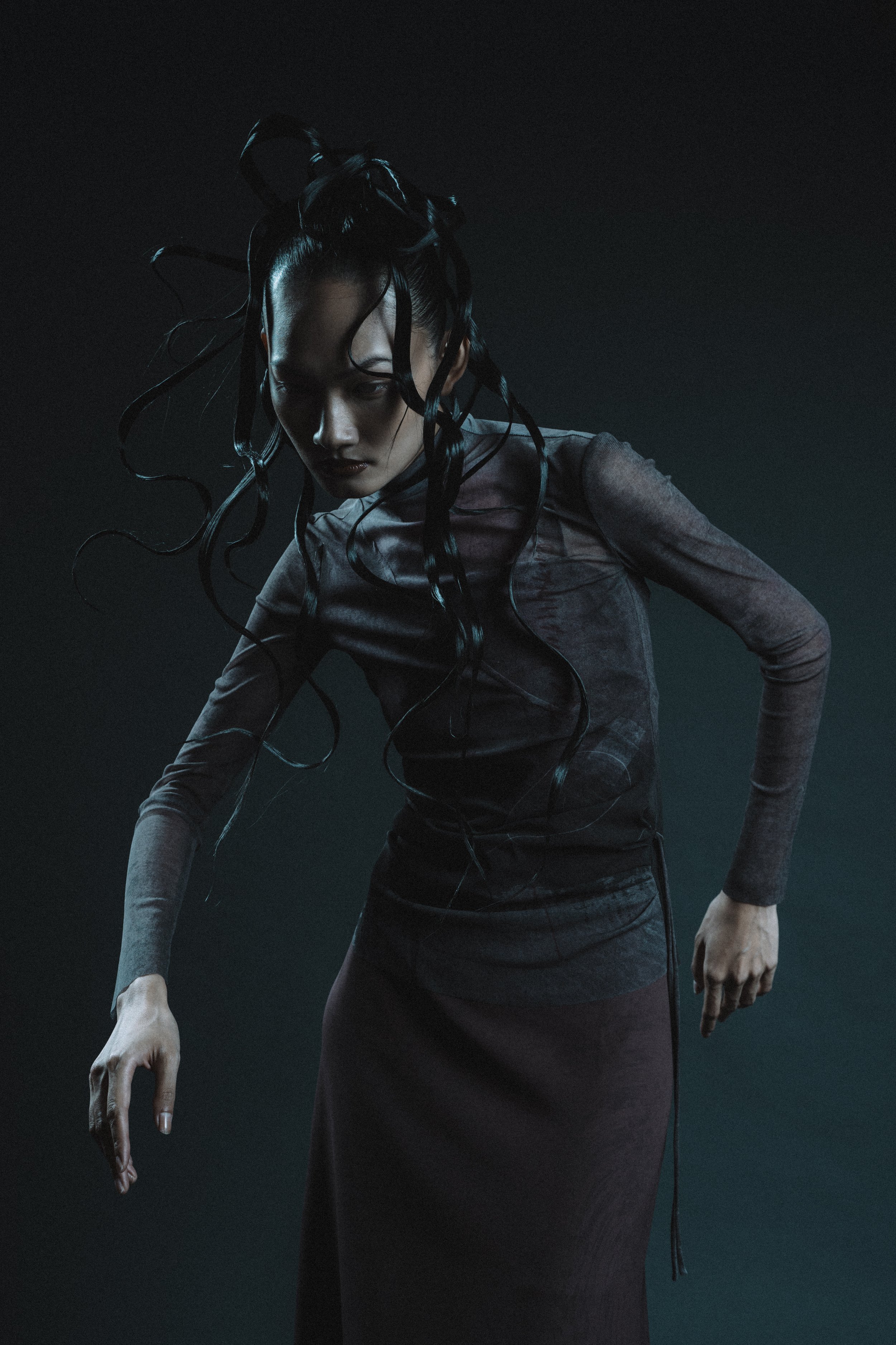 A woman with long, curly black hair, wearing a dark, shiny top and a long skirt, standing against a dark background with teal lighting.