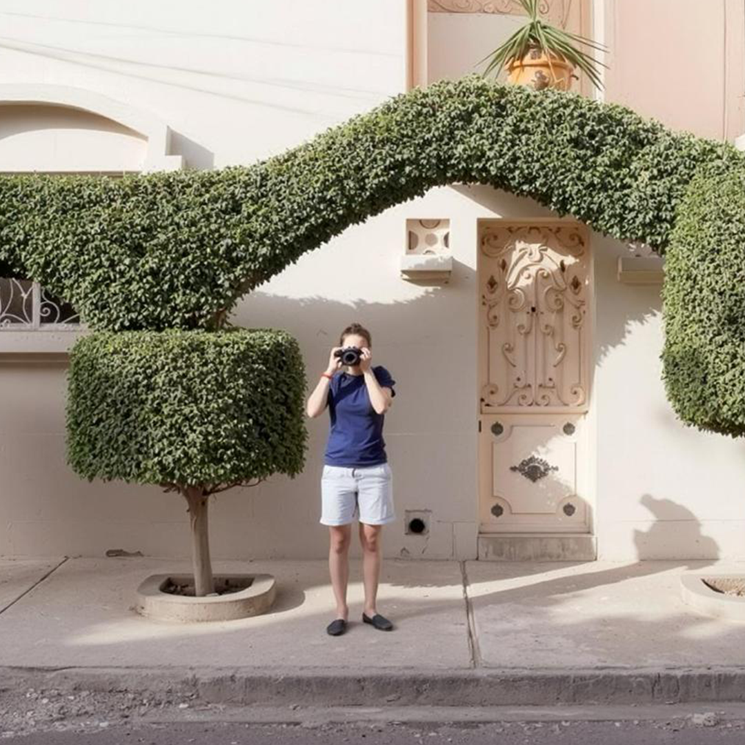 A person taking a photo with a camera in front of a decorative house facade with a house door, a small tree, and trimmed hedge