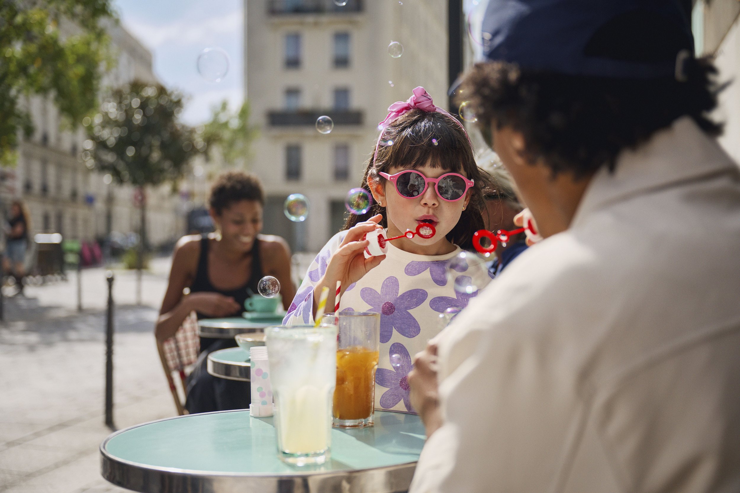 A young girl with dark hair, wearing pink sunglasses and a purple flower-patterned shirt, blowing bubbles at an outdoor cafe table. Two other people are seated at the table, and a woman with curly hair is in the background, also enjoying her time. Th