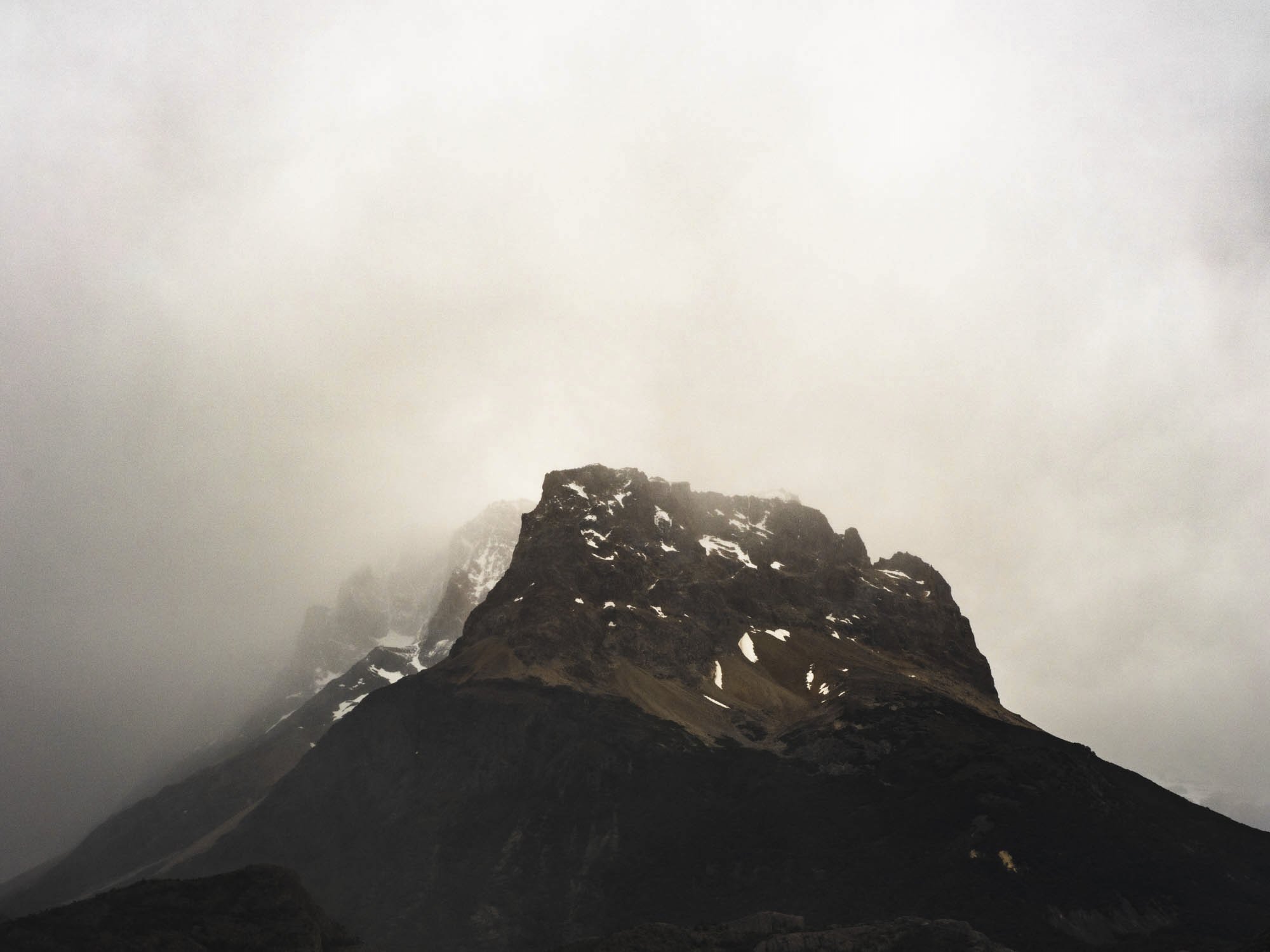 A dark mountain peak with snow patches, surrounded by foggy, overcast sky.