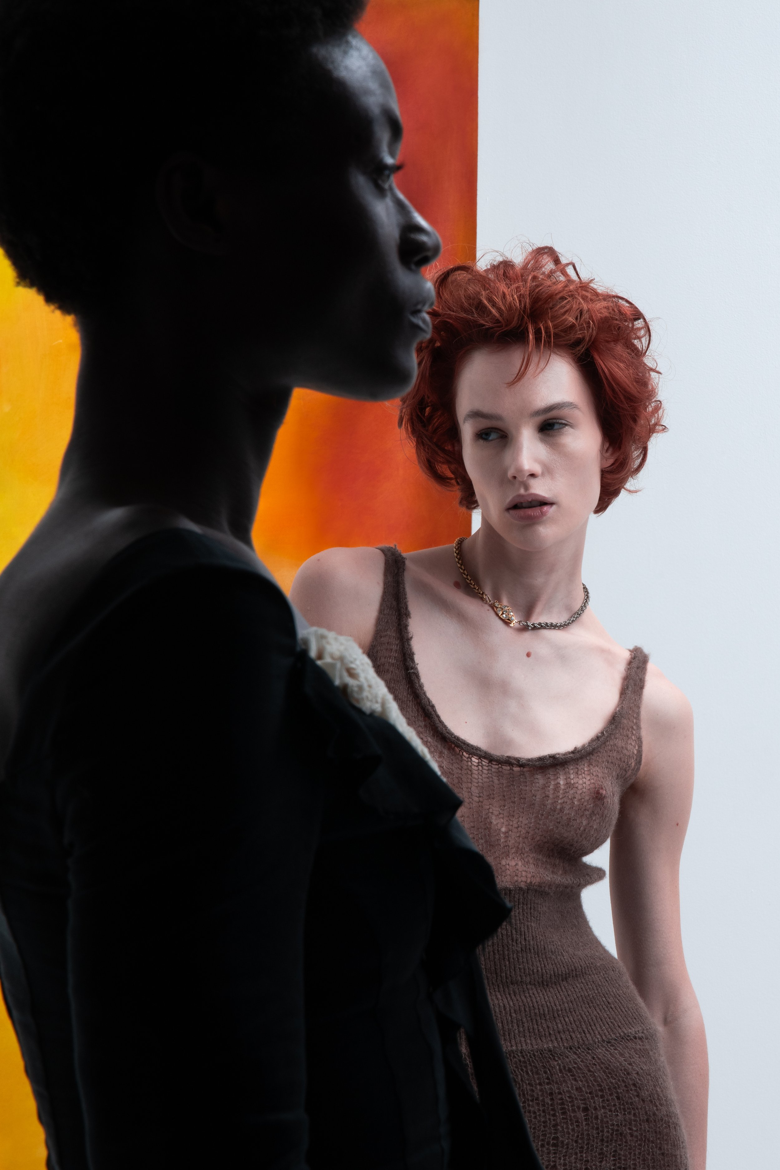 A woman with red curly hair wearing a brown tank top and a gold necklace looks at a woman with dark hair in a black outfit standing in front of a colorful orange and yellow background.