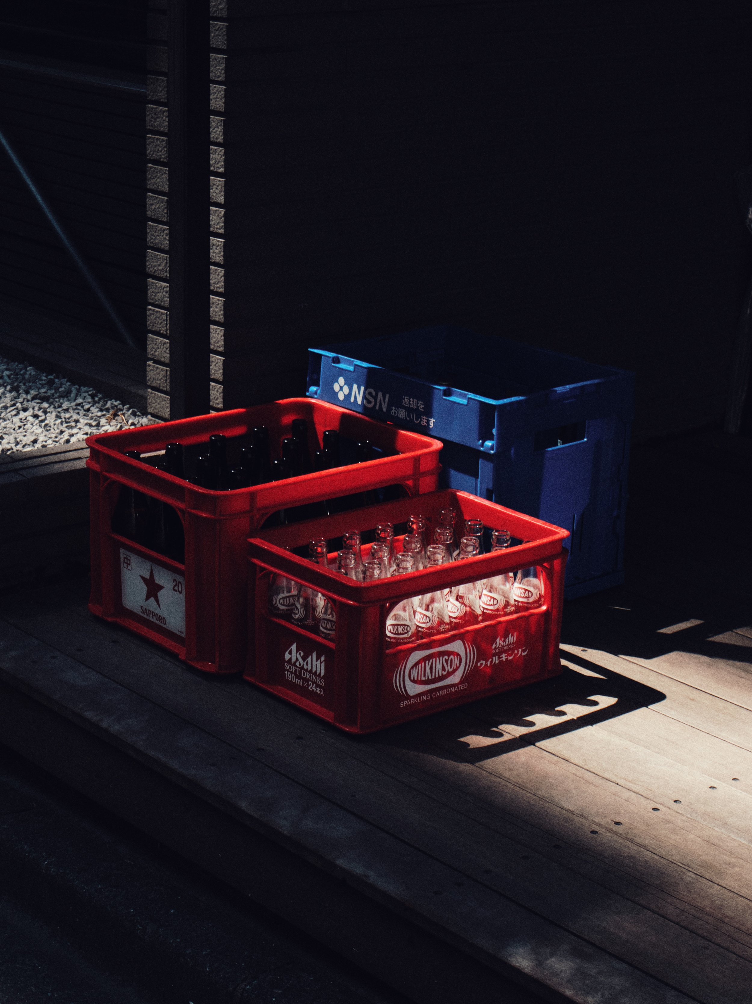 Three plastic crates of bottles, two red and one blue, stacked against a dark background on a wooden deck.
