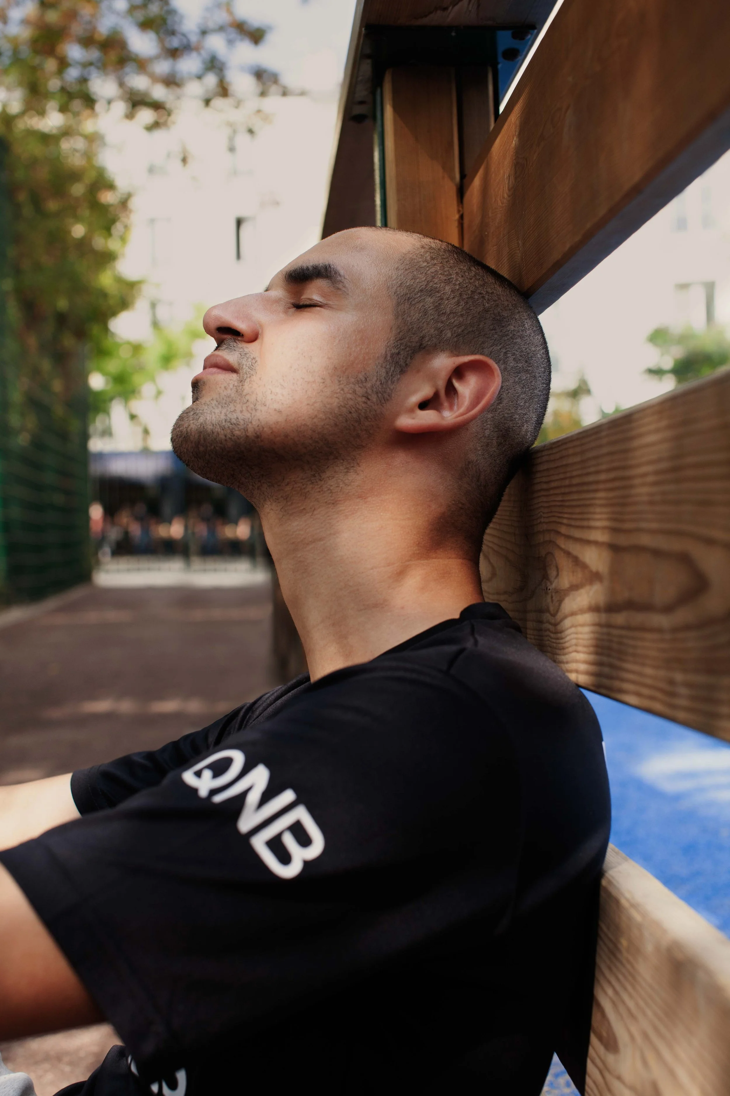 A young man with short hair and stubble relaxing with his eyes closed, leaning against a wooden fence, outdoors during daytime.