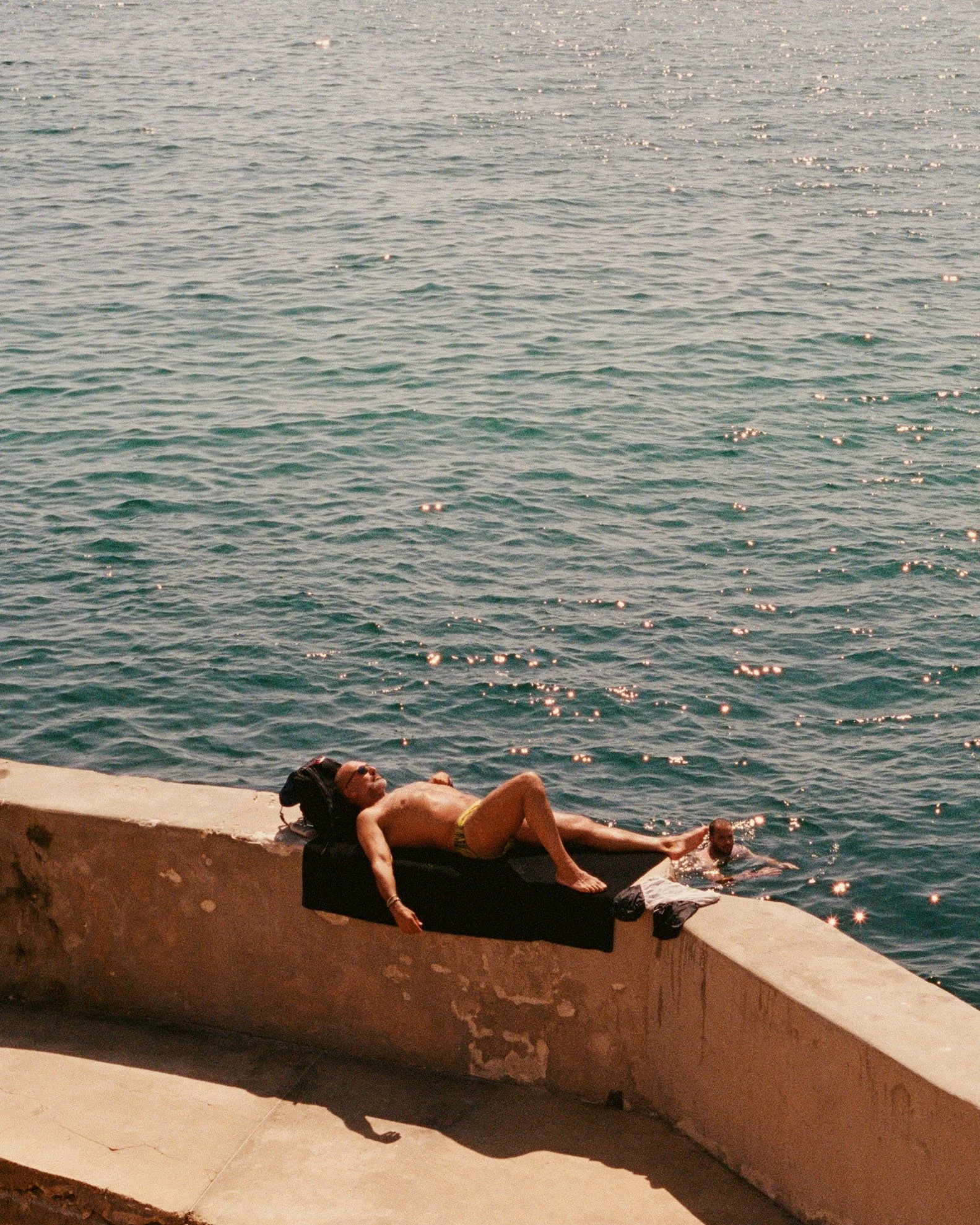 A man lying on a black lounge chair on a concrete pier, sunbathing near the water, with a person swimming in the water nearby.