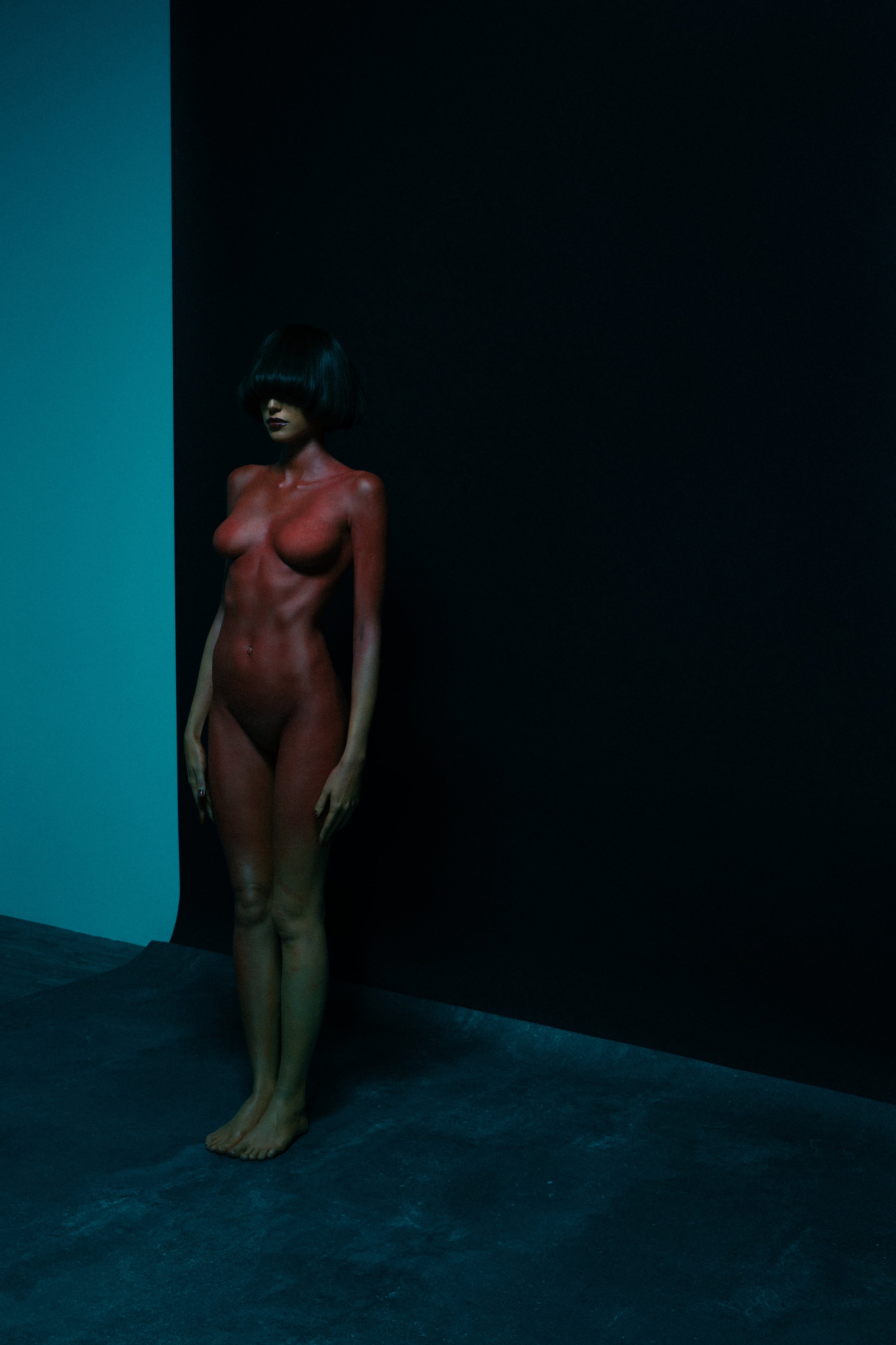 A woman with a black bob haircut and minimal clothing stands against a dark, contrasting background, partially illuminated by soft lighting.