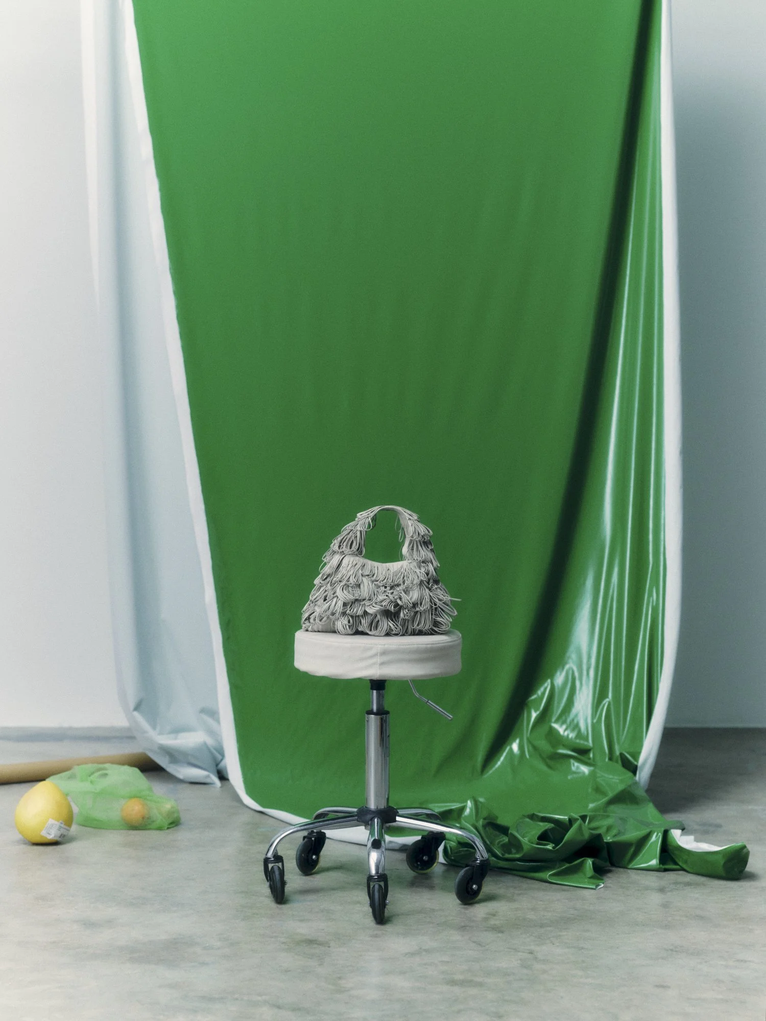 A green fabric backdrop with white sides and scattered objects including lemons, a green mesh bag, and a yellow item on the floor, with a chair in front.