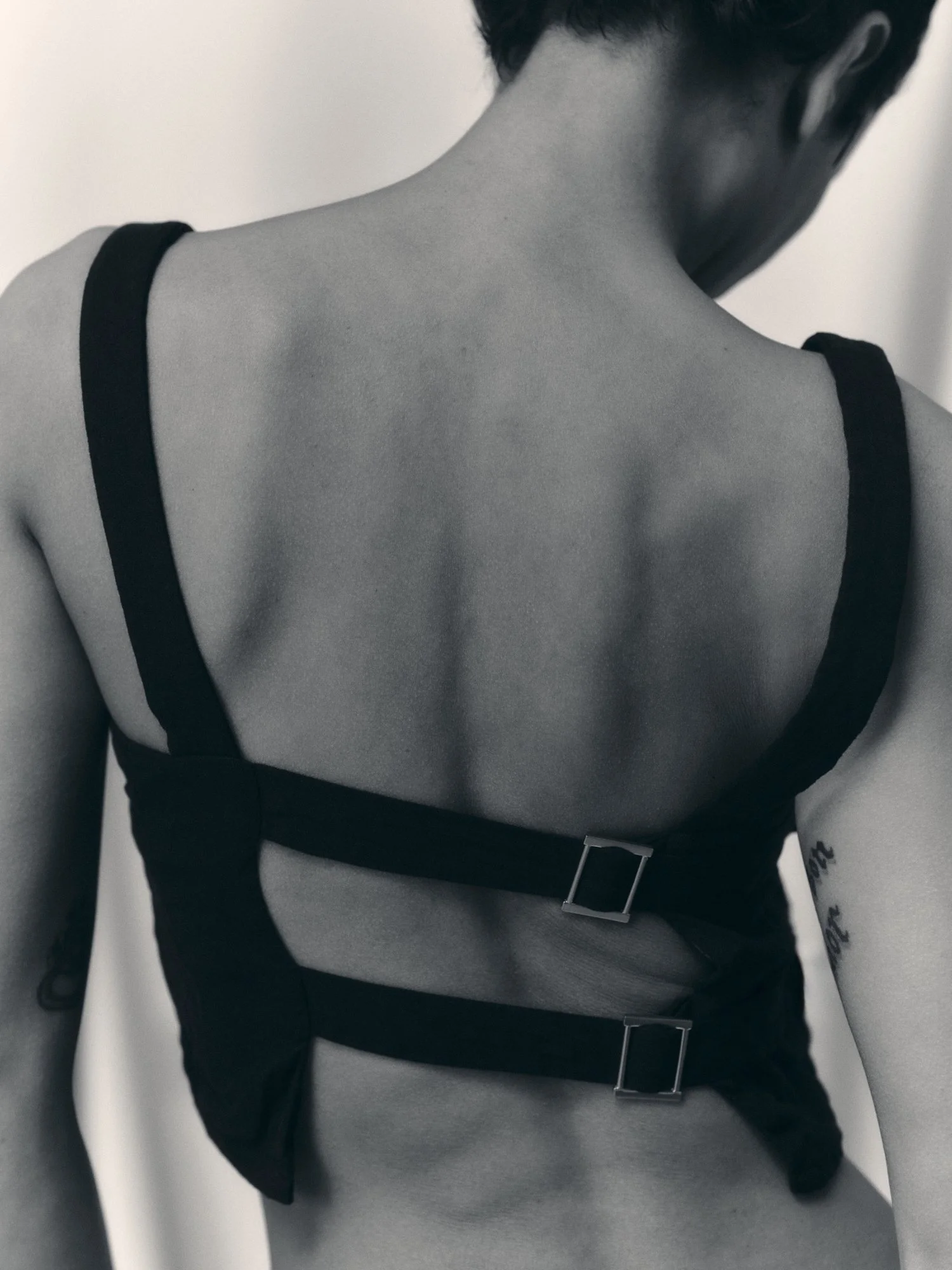 A person with short dark hair, wearing a black tank top, black pants, and a black harness with silver buckles, has a tattoo on their right forearm and is standing against a plain wall.