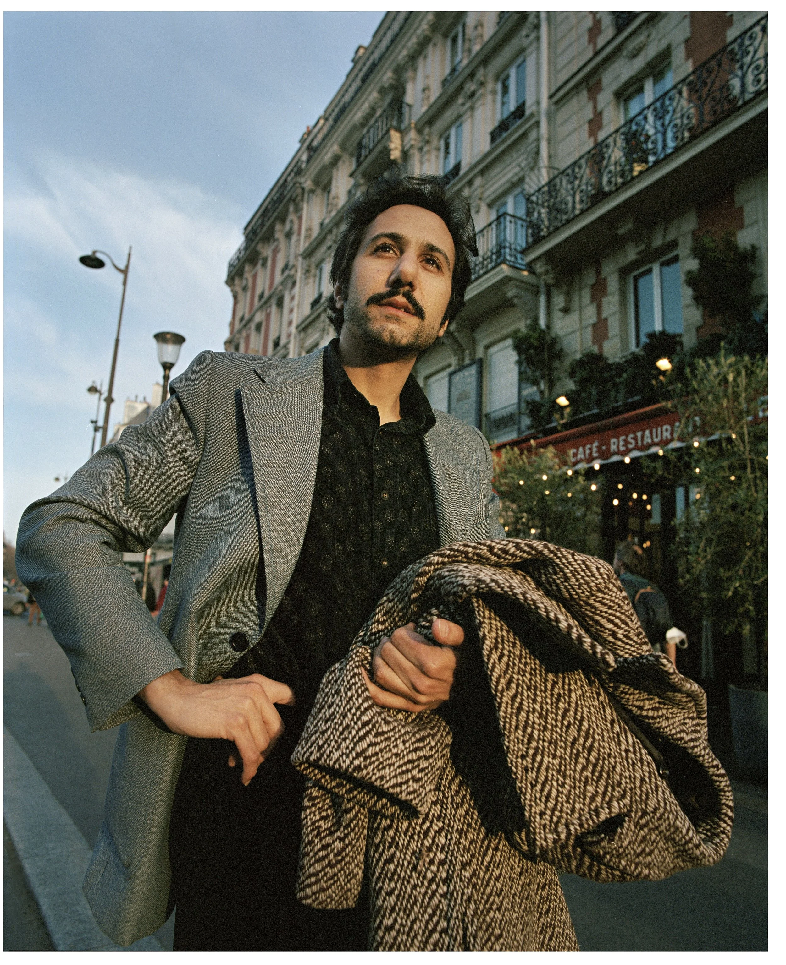 A man with dark hair and a mustache stands on a city street holding a coat, wearing a gray blazer and black shirt, with buildings and a café in the background.